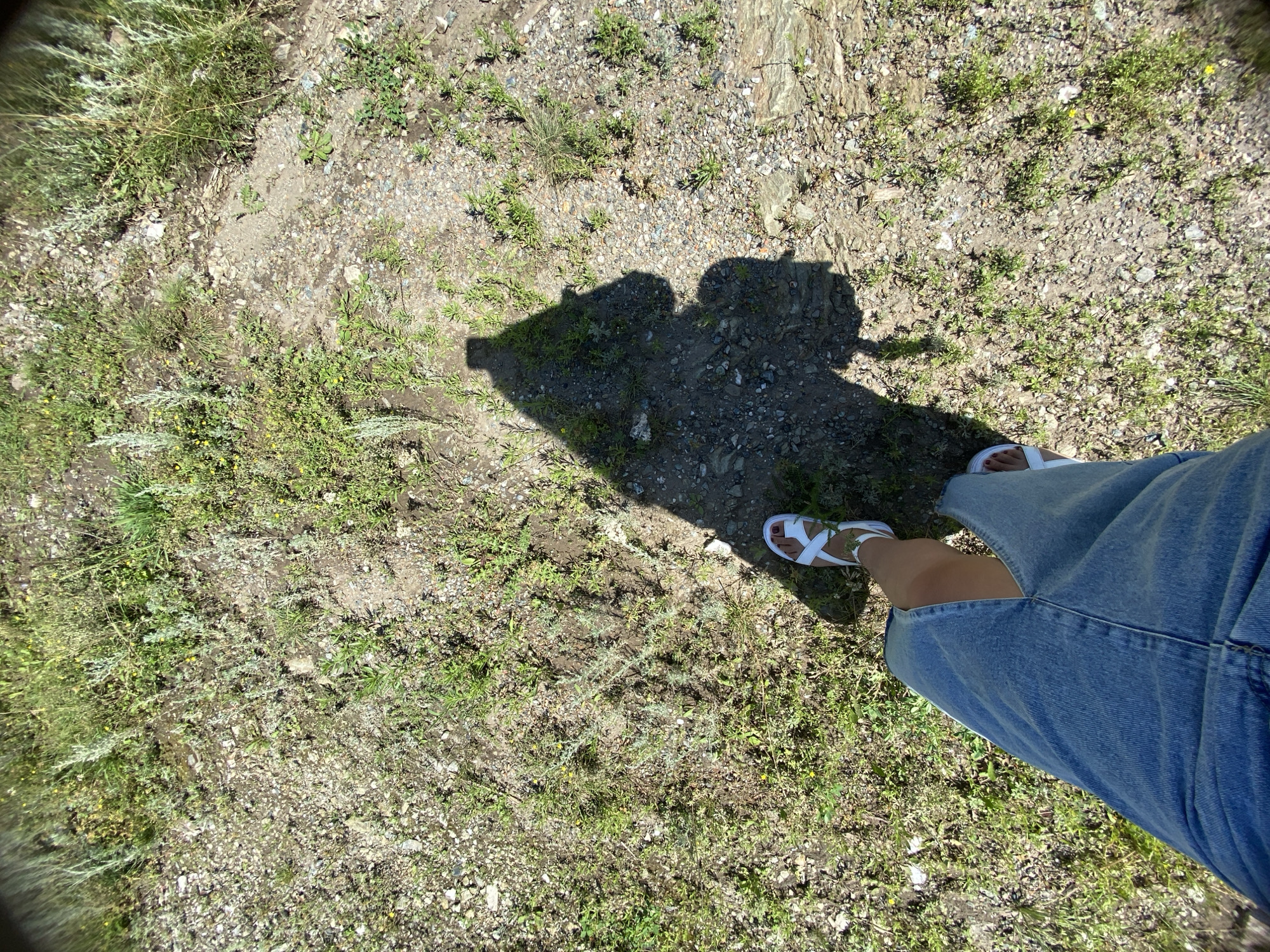 This is a first-person, downward-facing, wide-angle shot of a person standing on gravelly ground covered with sparse grass and small plants. The location is Yuldybayevo, Russia. The person is wearing a denim skirt and white sandals. A shadow of the person, cast by a bright sun, is visible on the ground. The shadow suggests the person is holding a camera or phone at arm's length. The overall environment appears to be an outdoor, possibly rural or semi-rural setting, with dry, rocky soil and green vegetation.