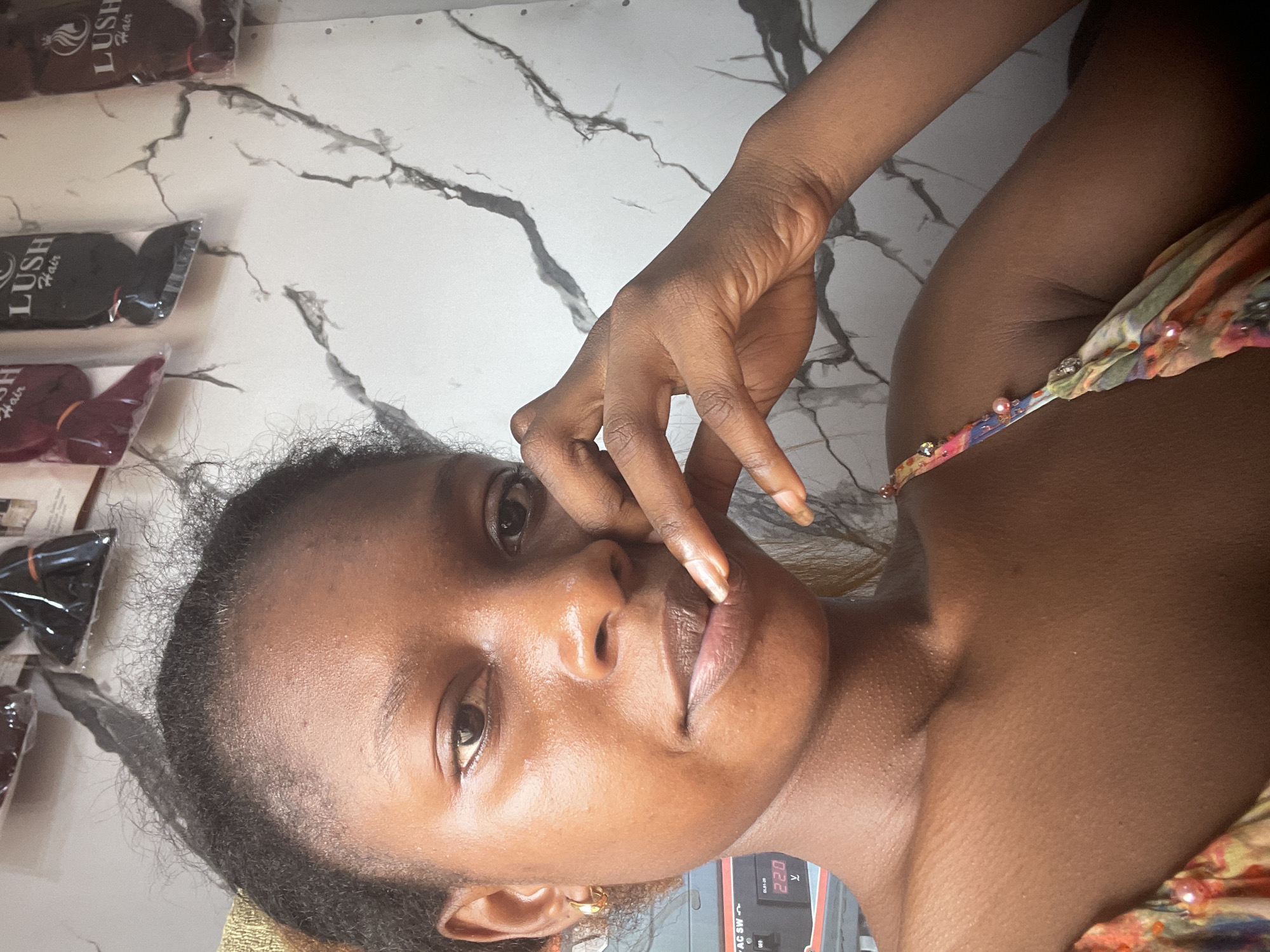 A close-up selfie captures a young Black woman with dark skin and natural hair posed indoors. Her right hand is raised to her face, with her fingers gently touching her lips. She wears a gold hoop earring in her left ear and a colorful dress with beaded straps. Behind her, shelves display packaged hair extensions, and a marble-patterned wall serves as the backdrop. A digital display unit is visible on the left side of the frame. The setting appears to be a shop in Lafia, Nigeria, possibly a hair salon or beauty supply store.