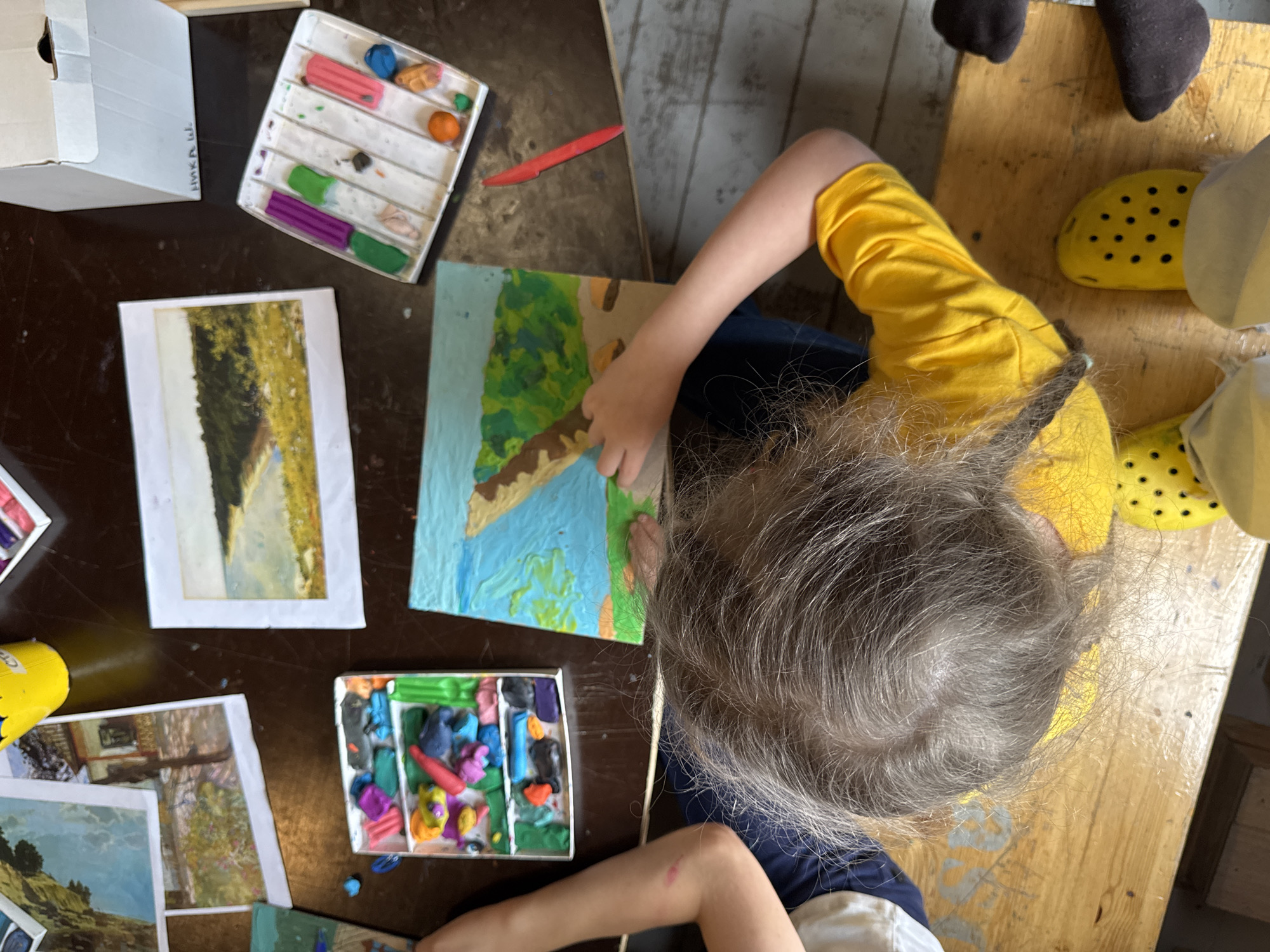 The image captures a top-down view of a table cluttered with art supplies, likely in a home setting. A child, seen from the shoulders down, is engrossed in creating art with colorful modeling clay. The child's hair is long and graying, and they are wearing a bright yellow shirt. On the table, there are several trays of modeling clay in various colors, including pink, green, purple, orange, and blue. There is also a small painting of a landscape, and what appears to be a red plastic tool. Another child's arm is visible on the left side of the frame, also engaged in artistic activity. The overall environment suggests a creative and playful atmosphere. The location is not specified, and it is not possible to determine if it is in Moscow, Russia.