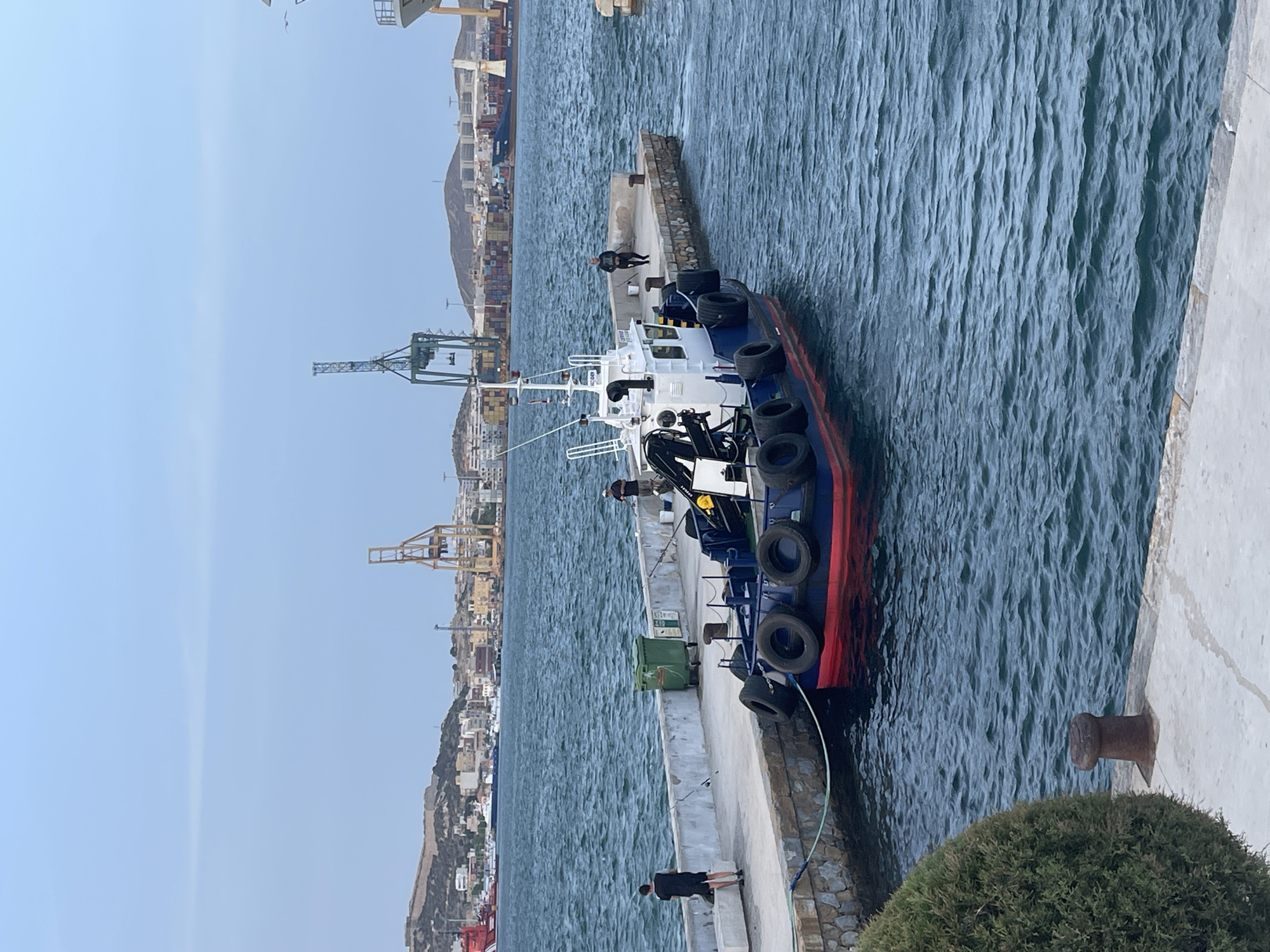 In Cartagena, Spain, a small, blue and red boat with a white superstructure is docked at a concrete pier. Several black fenders are attached to the boat's side. Two individuals stand on the pier near the boat. One, wearing a black t-shirt and shorts, is facing away from the camera and towards the water, appearing to be fishing. Another person, in a dark shirt and pants, stands further down the pier, also facing away. A green trash receptacle is present on the pier. In the background, the city of Cartagena is visible with buildings and hills, and several large cranes dominate the skyline. The water is choppy, and the sky is overcast.