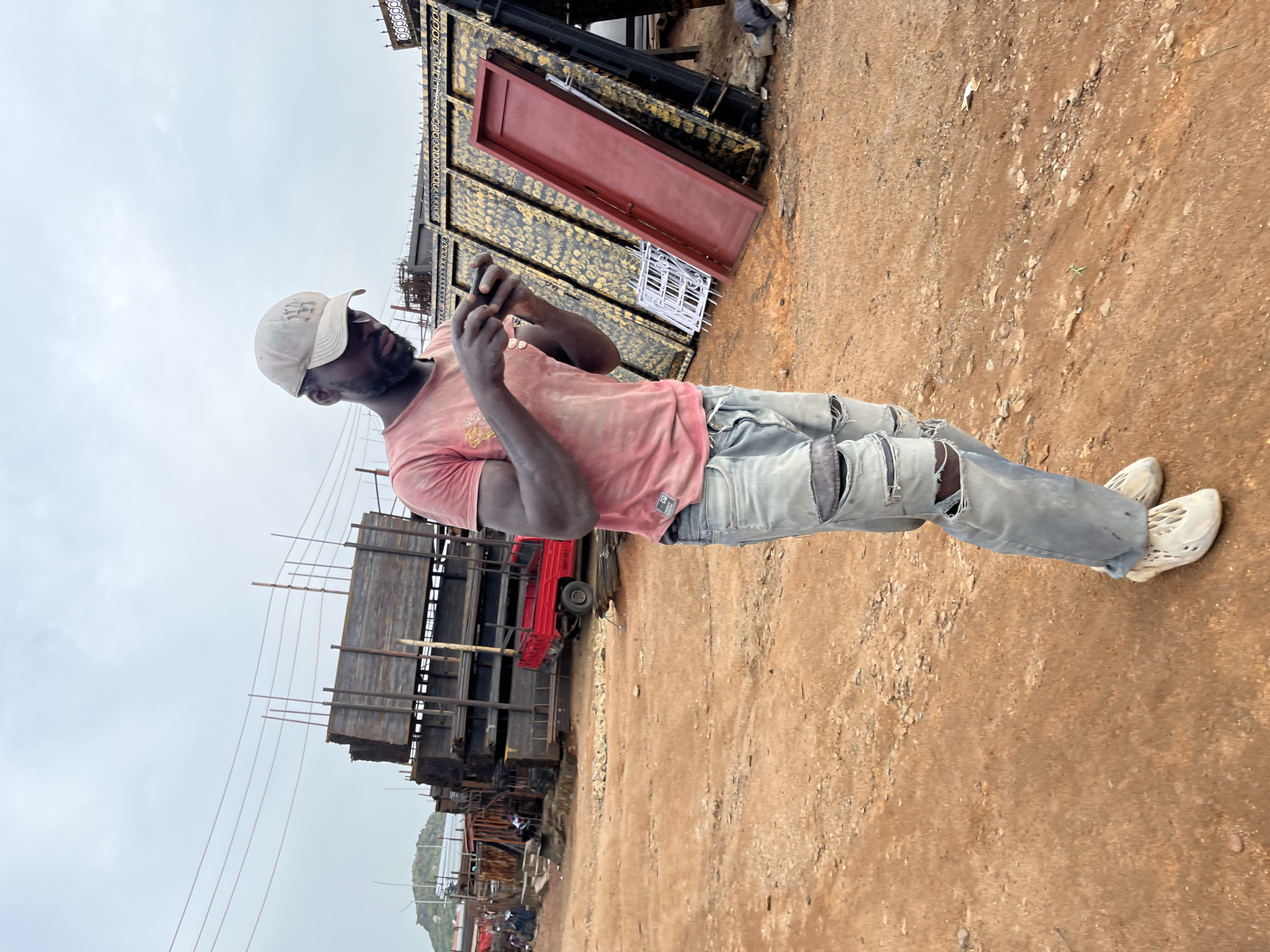A man wearing a light-colored baseball cap, a pink t-shirt, and ripped light-blue jeans stands in a dusty outdoor area. He is looking down at his hands, which are holding a dark-colored mobile phone, suggesting he is using it. The man is positioned in front of a large stack of what appear to be metal gates or structures, some with red accents. Behind these, more industrial materials are visible, including what looks like a red truck. In the background, there are more stacks of metal, scaffolding, and a cloudy sky. The ground is predominantly dirt and gravel, indicative of a construction site or storage yard. The overall scene suggests a working environment, possibly a fabrication or construction material yard. The lighting is diffused, consistent with an overcast day.