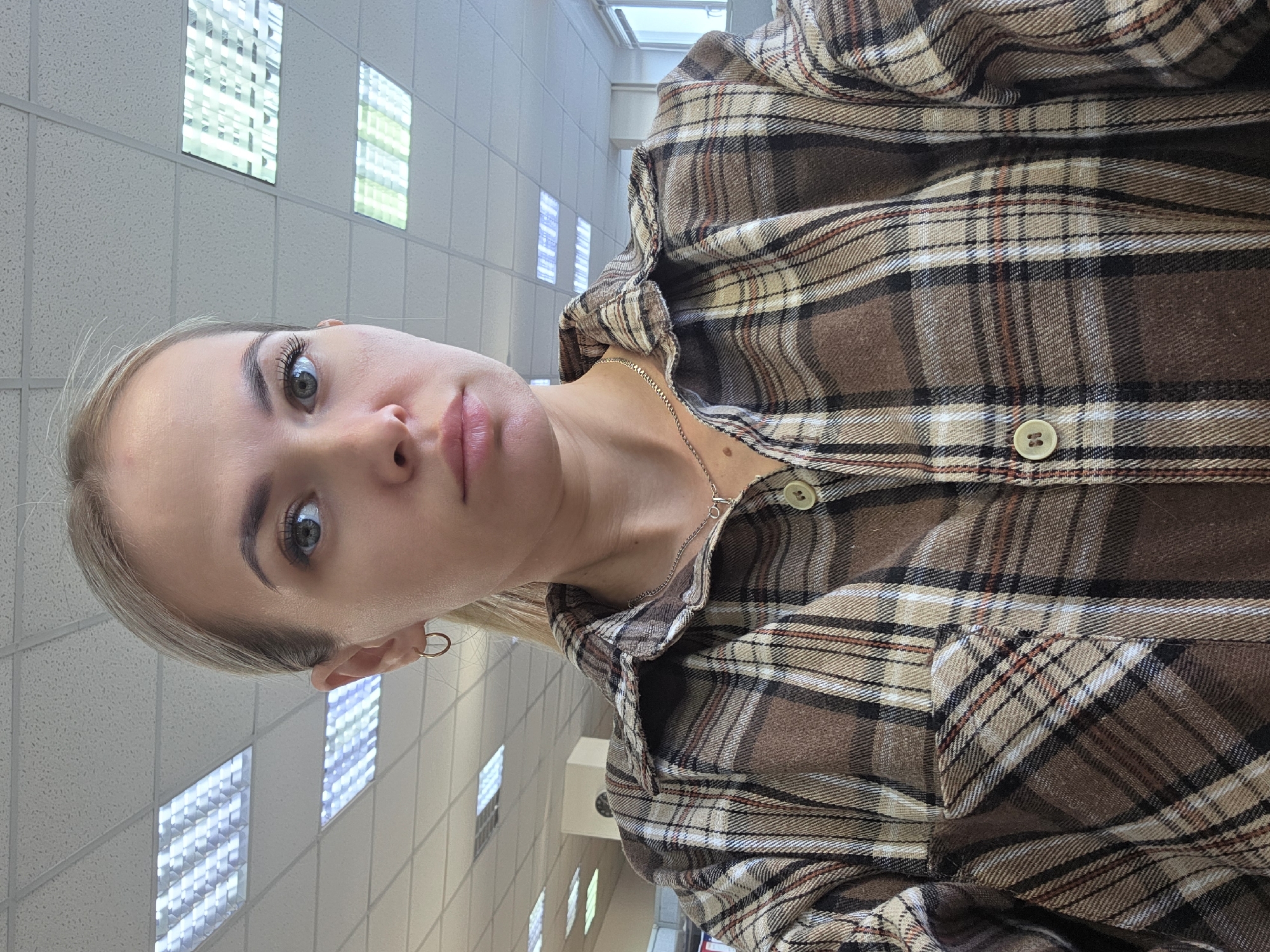 A selfie of a fair-skinned woman with light blonde hair is oriented vertically. She is wearing a brown, tan, and white plaid flannel shirt with two visible buttons, a delicate silver chain necklace, and small hoop earrings. The background shows a ceiling with recessed lighting arranged in a grid pattern, suggesting an interior setting. The location information is not discernible from the provided image.