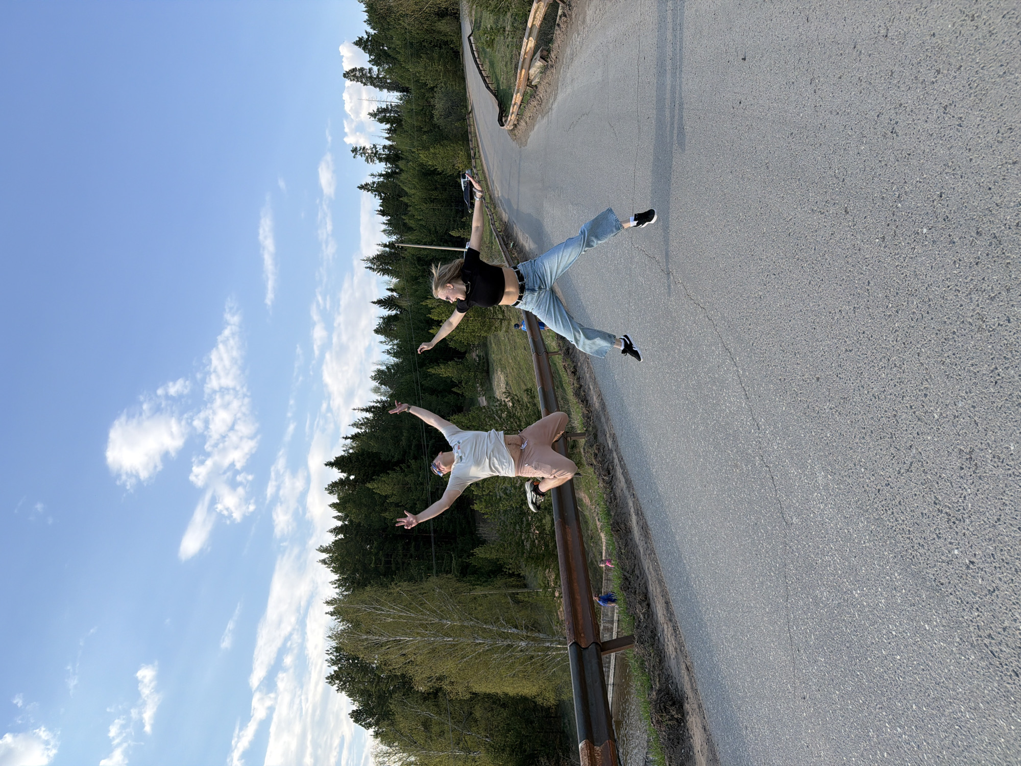Two individuals are captured in mid-air on what appears to be a paved road with a metal guardrail. The road winds slightly, with a backdrop of dense green trees and a blue sky dotted with clouds. The top figure is a person with light hair, wearing a black top and blue jeans, appearing to jump. The lower figure is a person wearing a light-colored shirt, pink shorts, and sunglasses, positioned on the guardrail. The location is likely outdoors, possibly on the outskirts of the city of Kamennoye, Russia, given the presence of trees and the road.