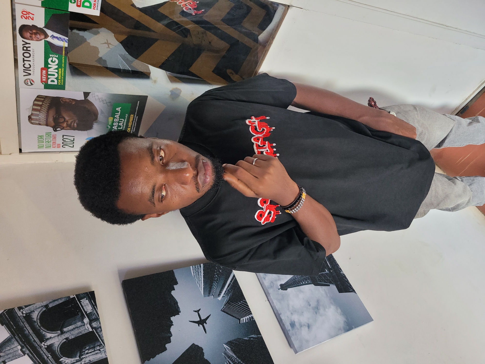 The image shows a young Black man with dark hair and a beard, wearing a black t-shirt with red lettering and light grey pants. He is standing in a room with white walls adorned with framed pictures. The pictures depict architectural landmarks and a plane flying between buildings. To the right of the man, there are posters with political campaign imagery, including photos of candidates and campaign slogans. The overall setting suggests an indoor space, possibly a residence or a small office, decorated with personal and political memorabilia. The man is looking down and to his left, with his right hand touching his chin. His left hand is in his pocket. He has a ring on his left ring finger and bracelets on his right wrist. One of the posters features the name "KEVIN DUNG" and "FOR MEMBER HOUSE OF ASSEMBLY JOS-SOUTH CONSTITUENCY". Another poster shows a man in traditional attire with the text "BAS BALA LAU YAN BRRN (LAU)" and "FOR MEMBER". The presence of these posters indicates that the location is likely Jos, Nigeria, as they relate to local political elections. The room appears to be lit by artificial light.