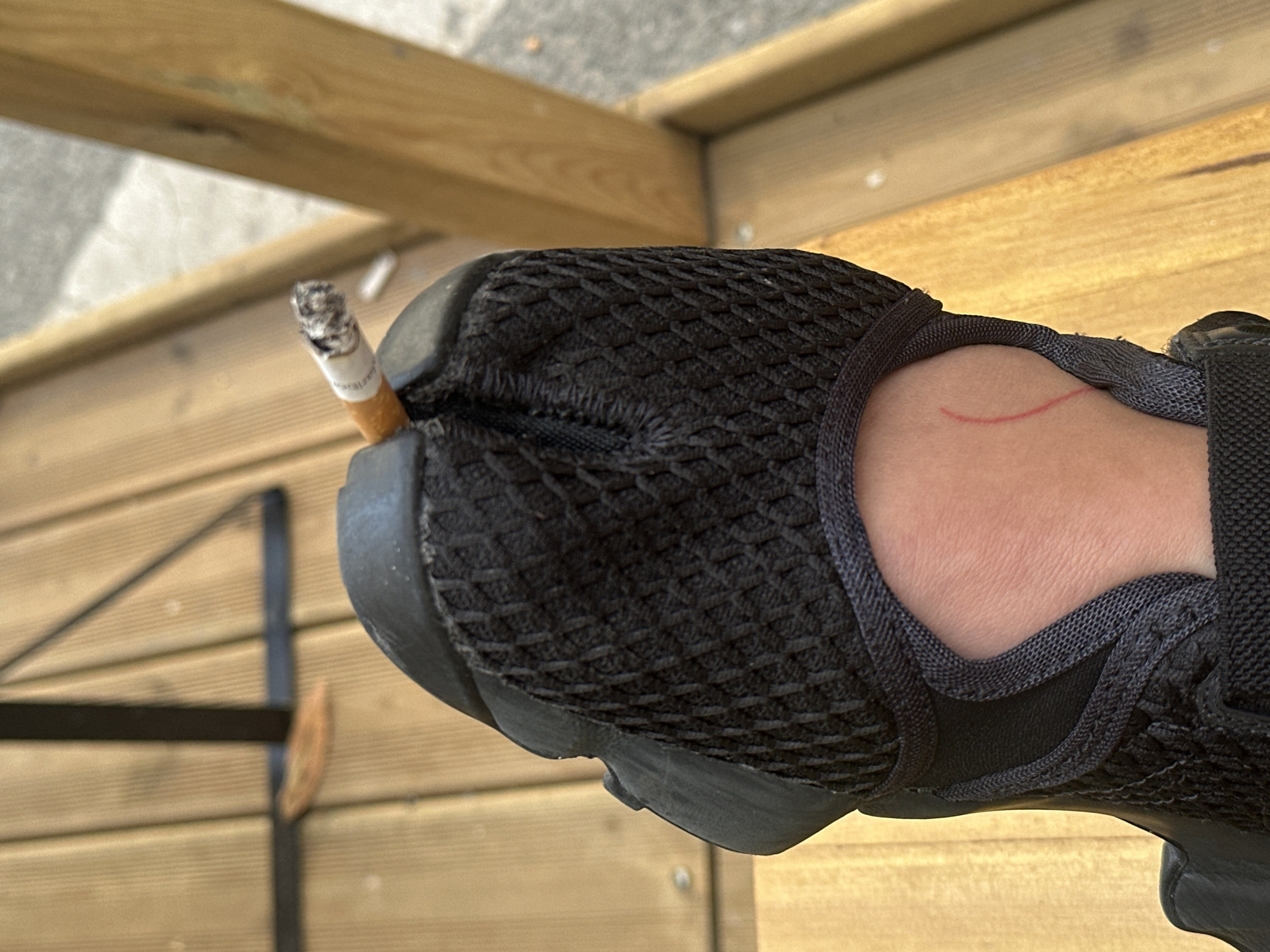 A close-up photograph shows a black athletic shoe with a lit cigarette wedged into the toe. The shoe is being worn, evidenced by the exposed ankle and foot visible in the frame. A red mark is visible on the wearer's ankle. The background features wooden planks, potentially a deck or outdoor seating area. A black metal object, possibly part of outdoor furniture, is also partially visible. The image does not provide sufficient information to determine the location within Saint-Ouen-sur-Seine, France.