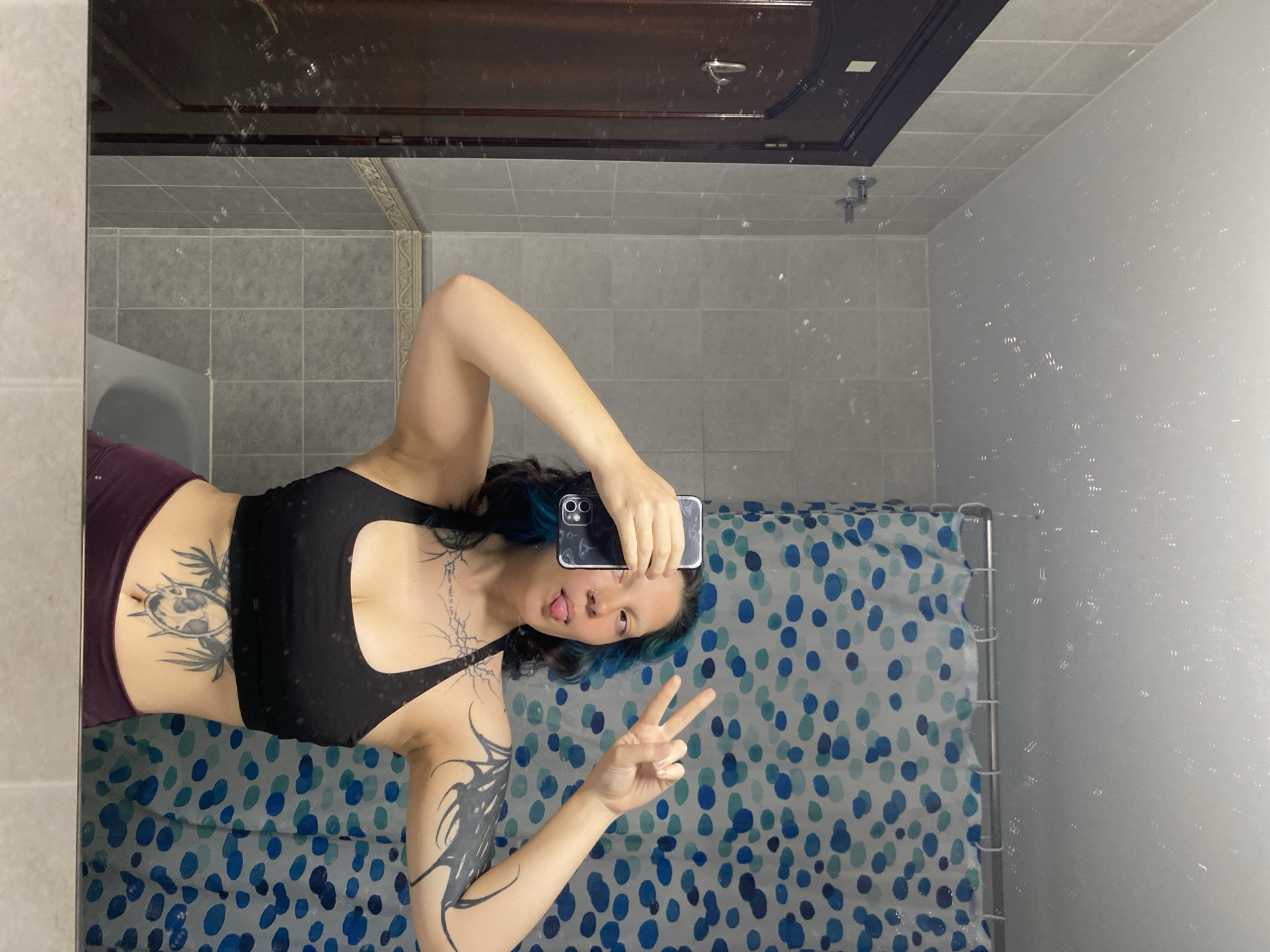 A person is taking a selfie in a bathroom. The person is wearing a black sports bra and dark purple pants, and has blue-tinged hair, tattoos on their arms and stomach, and is holding up two fingers while sticking out their tongue. The bathroom has a shower curtain with a blue and white polka dot pattern, grey tiled walls, and a wooden door. The reflection in the mirror shows a blurry image of the bathroom. It's impossible to determine whether this image was taken in the city of Gijón/xixón, Spain, as there are no identifying landmarks or features visible.