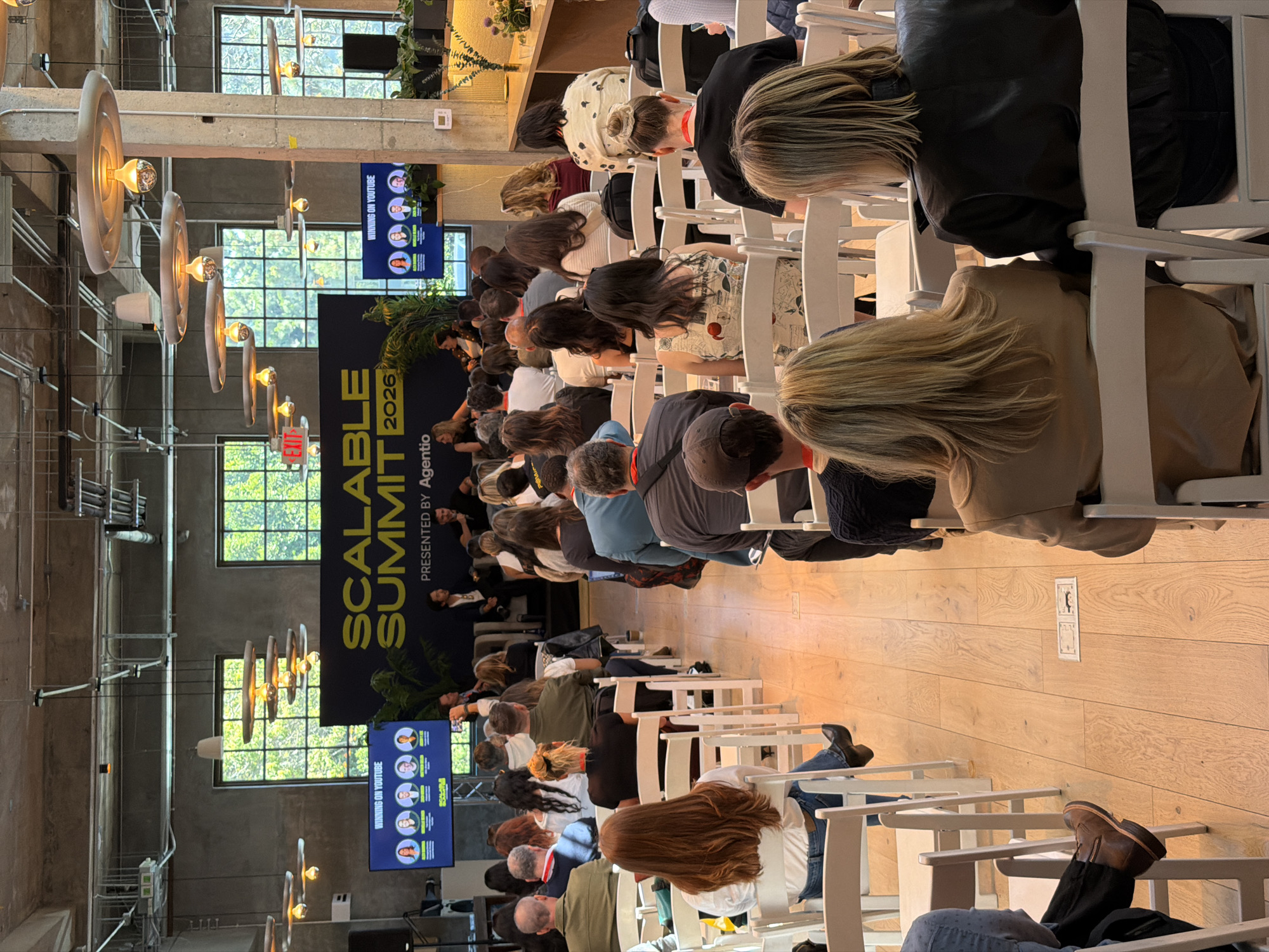 The image shows a gathering of people seated in white chairs, attending what appears to be a conference or presentation. A large backdrop displays the words "SCALABLE SUMMIT 2026," along with smaller displays showing the text "WINNING ON YOUTUBE". The scene is lit by industrial-style hanging lights. The location is in Los Angeles, United States of America.