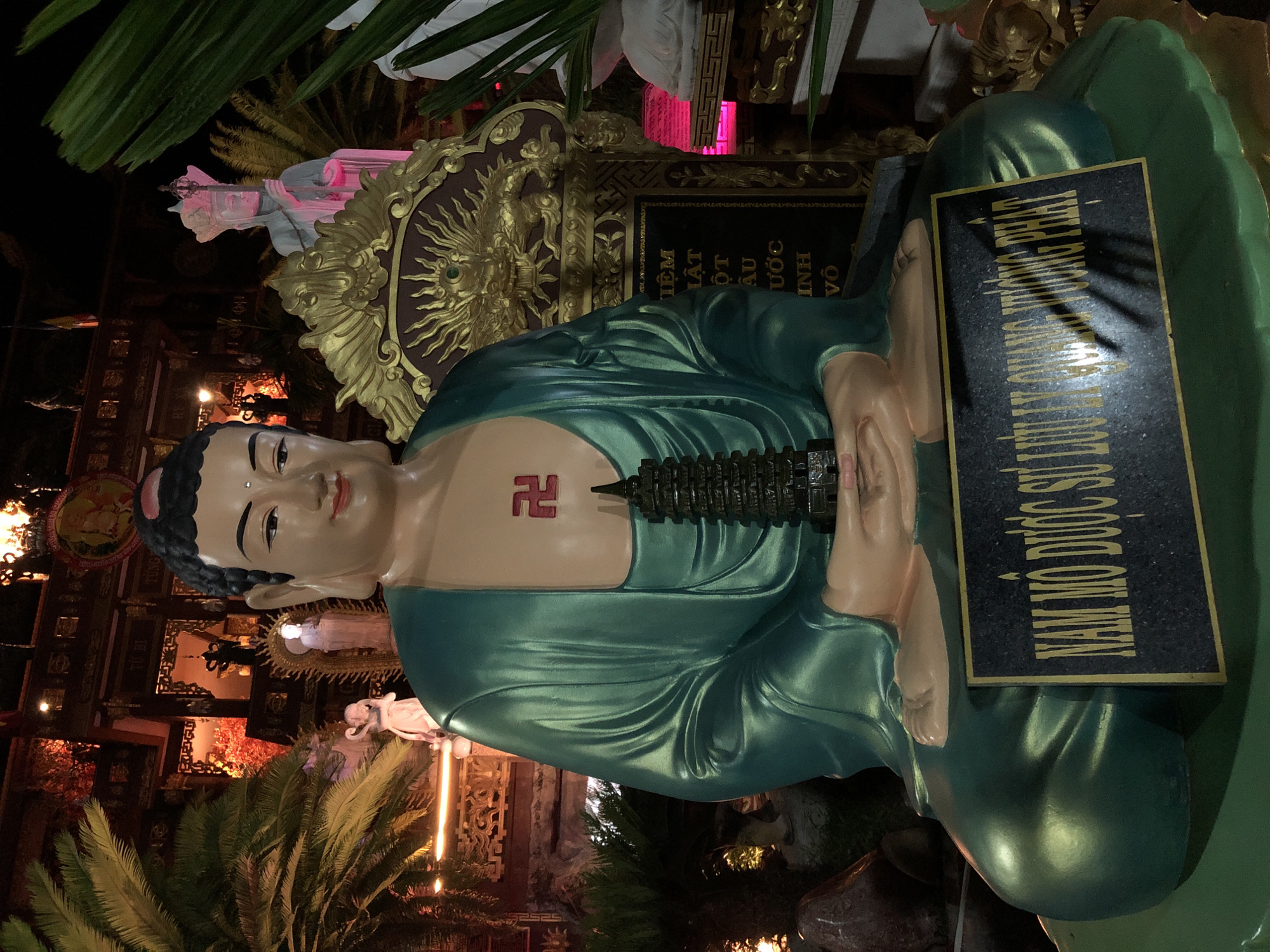 The image shows a statue of a Buddha in a sitting position, adorned with a green robe. The statue is placed in a dimly lit setting, possibly outdoors at night, with vibrant greenery and architectural elements in the background, suggesting a temple or a garden in Mũi Né, Vietnam. A prominent detail on the Buddha's chest is a red swastika symbol, which is an ancient symbol of good fortune and well-being in Buddhism and other Eastern religions. There is also a small pagoda-like object placed vertically on the statue's chest.
A plaque with gold lettering is attached to the statue's base. The text on the plaque appears to be in Vietnamese. The visible portion reads "NAM MO DUC SU LUULTUONG PHAT" and part of another line below. The statue is illuminated by warm, artificial lights, creating a mystical ambiance. The overall scene conveys a sense of serenity and spiritual devotion.