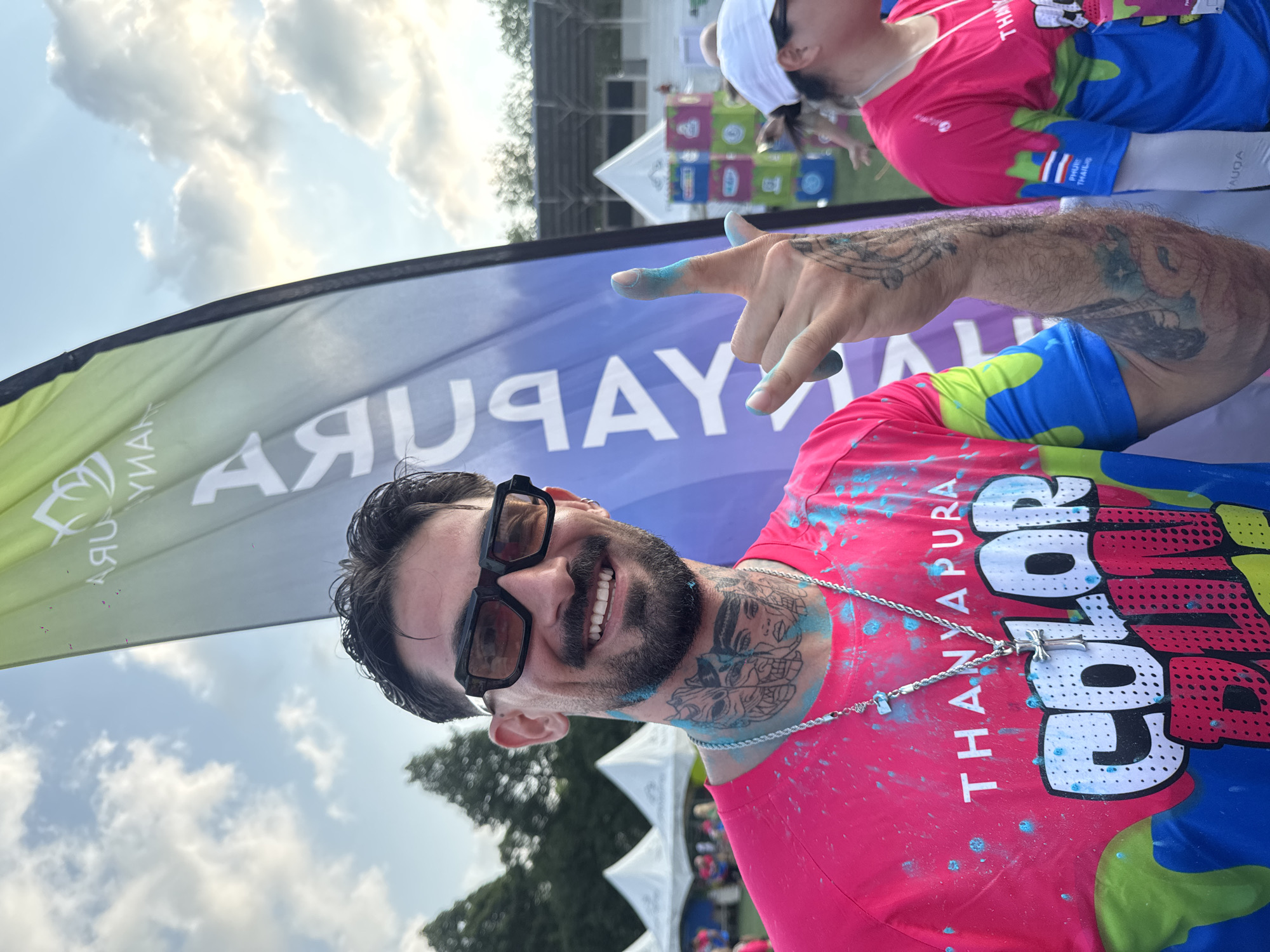 The image depicts a man with a beard, wearing glasses and a pink T-shirt with the words "Color Thanayapura" splattered with blue paint. He is holding up two fingers covered in blue paint, indicating the peace sign. The man has tattoos on his arms and neck, and a silver chain with a cross pendant around his neck. Behind him is a banner with the word "Thanayapura" visible. In the background, there are other people, tents, and trees, suggesting the event is outdoors in Thep Krasatti, Thailand.