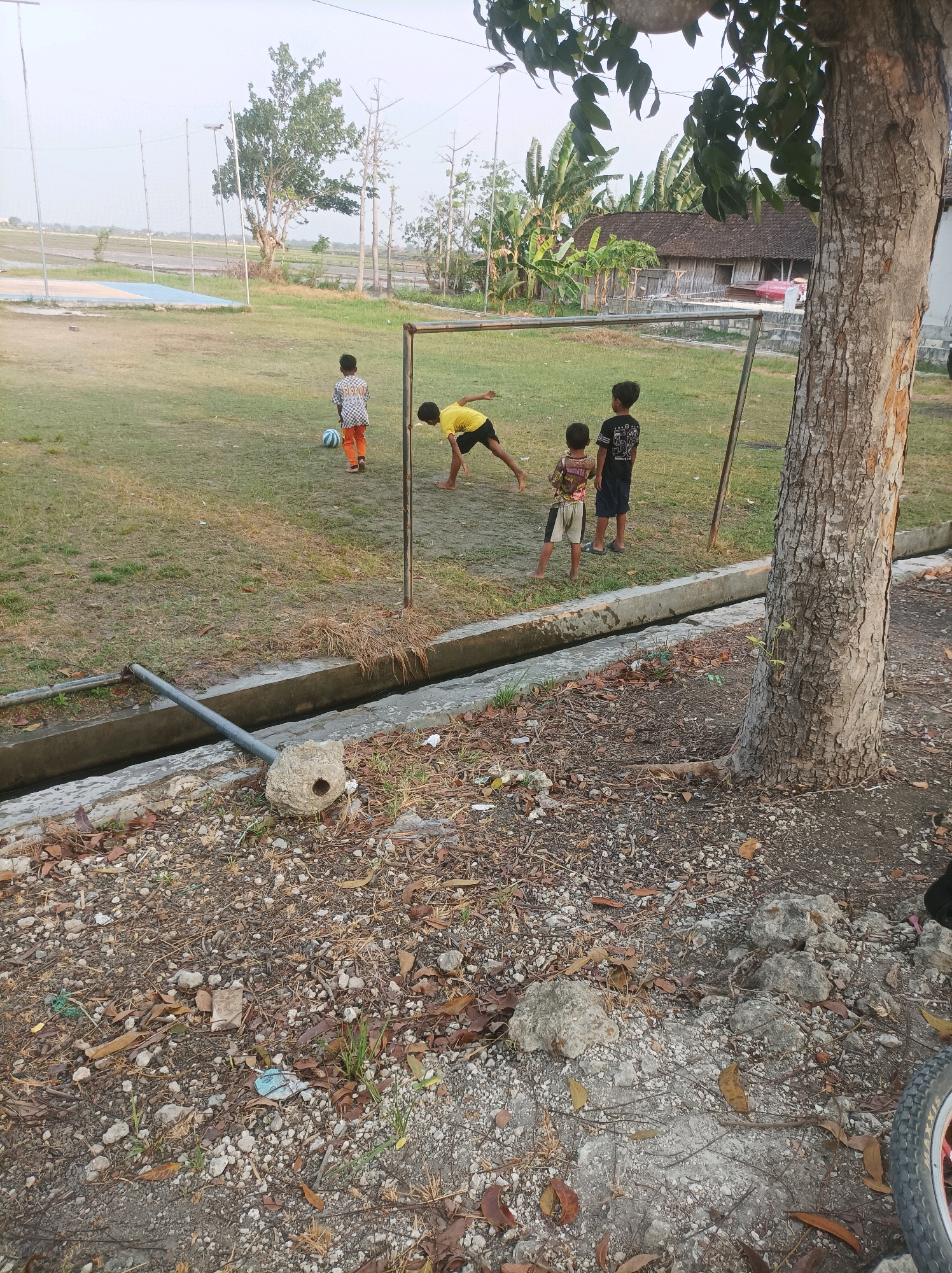 A group of four young boys are playing soccer in an outdoor grassy area with a goalpost. One boy in an orange and white checkered shirt is kicking a blue and white soccer ball. Another boy in a yellow shirt is bent over, possibly receiving a pass or about to kick the ball. Two other boys are standing near the goalpost, one wearing a patterned shirt and shorts, and the other in a black shirt and dark shorts. The environment appears to be a rural or semi-rural setting, with a wooden-roofed building visible in the background, surrounded by banana trees. A drainage ditch runs along the foreground, with a pipe extending into it and a wasp nest attached to its side. The ground is covered in dirt, gravel, and fallen leaves. The sky is overcast.