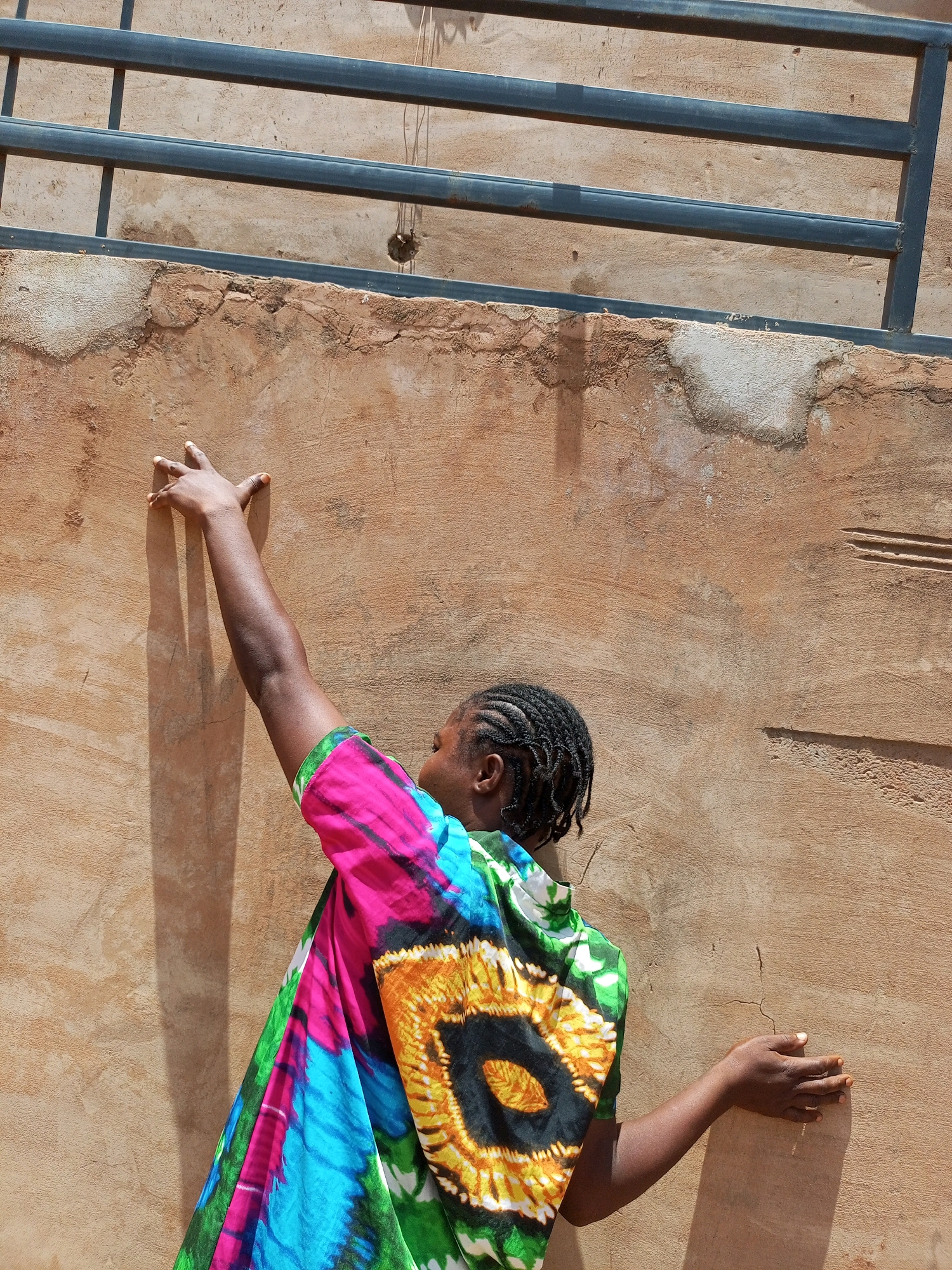 In Tempure, Nigeria, a person with braided dark hair is depicted from behind, facing a textured, ochre-colored wall. They are wearing a vibrant, tie-dye garment with a pattern featuring black, yellow, blue, and pink hues. The person's left arm is extended upwards, with their hand touching the wall. Their right arm is bent at the elbow, with their hand also touching the wall. Two horizontal dark metal bars are visible at the top of the frame, casting shadows on the wall. The wall itself shows signs of wear and tear, with cracks and patches of lighter plaster. The lighting suggests it is daytime, with distinct shadows cast by the person and the metal bars.