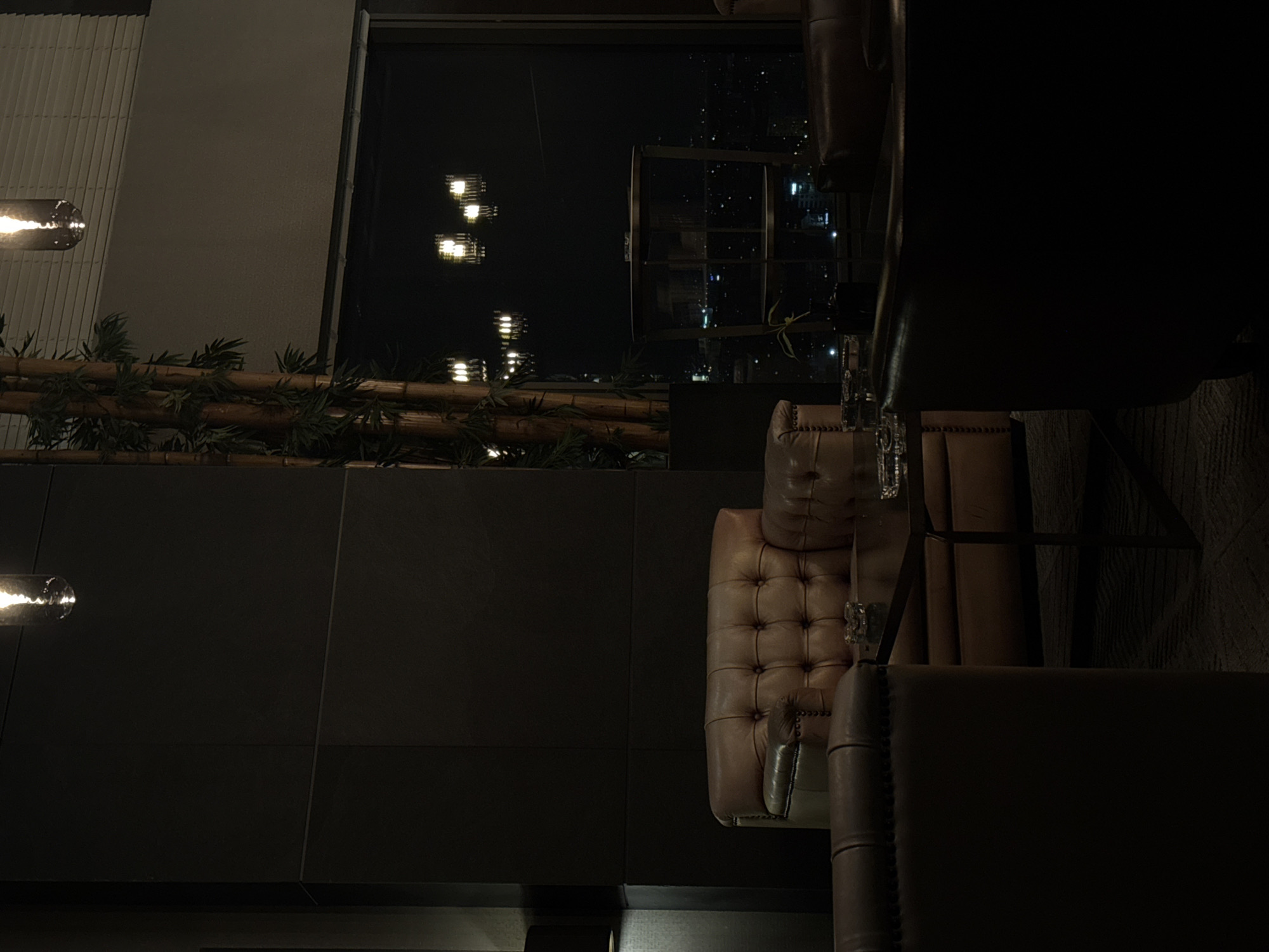 This is a photograph of a dimly lit, modern interior, likely a lounge or bar, with a large window overlooking a city at night. The architecture suggests a sophisticated setting, possibly in a high-rise building, providing a panoramic view of Sofia, Bulgaria.
In the foreground, a plush, brown leather Chesterfield sofa is prominently featured. Its tufted design and classic silhouette convey a sense of luxury and comfort. To the left of the sofa, another section of a similar brown leather armchair is visible, suggesting a seating arrangement designed for relaxation. A low, dark coffee table sits between these seating elements, with ashtrays on its surface, indicating a space where smoking may be permitted.
The background is dominated by a large window that reveals the nighttime cityscape of Sofia. Numerous lights from buildings and streetlights create a shimmering tapestry of urban illumination. The dark sky above is punctuated by the reflections of interior lights on the glass.
To the left of the window, a tall, decorative element resembling bamboo stalks stands vertically, adding an organic and textural contrast to the otherwise modern and man-made environment. Hanging light fixtures with a vintage or industrial design are visible both within the interior and reflected in the window, casting warm, ambient light.
The overall atmosphere is one of intimate, upscale leisure, designed to offer a comfortable and visually engaging experience with the city's nocturnal panorama. The scene appears devoid of any people, contributing to a sense of stillness and quietude.