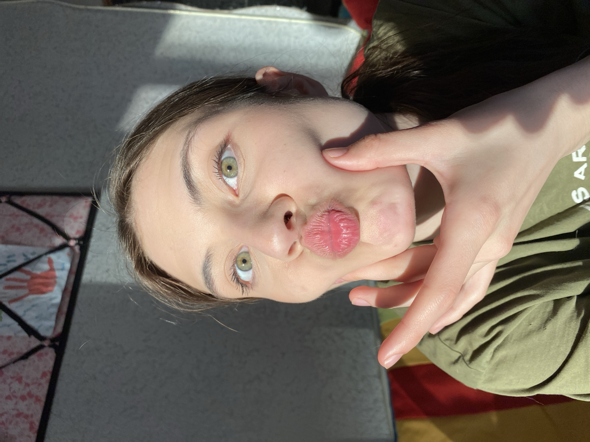 A young woman with green eyes is close to the camera, puckering her lips into a kiss. Her hand is positioned on her right cheek. She has light skin and brown hair. She wears a green shirt. The background appears to be a room with a bed and possibly a blanket with a handprint design. The lighting indicates a sunny day. The image was not taken in Votkinsk, Russia.