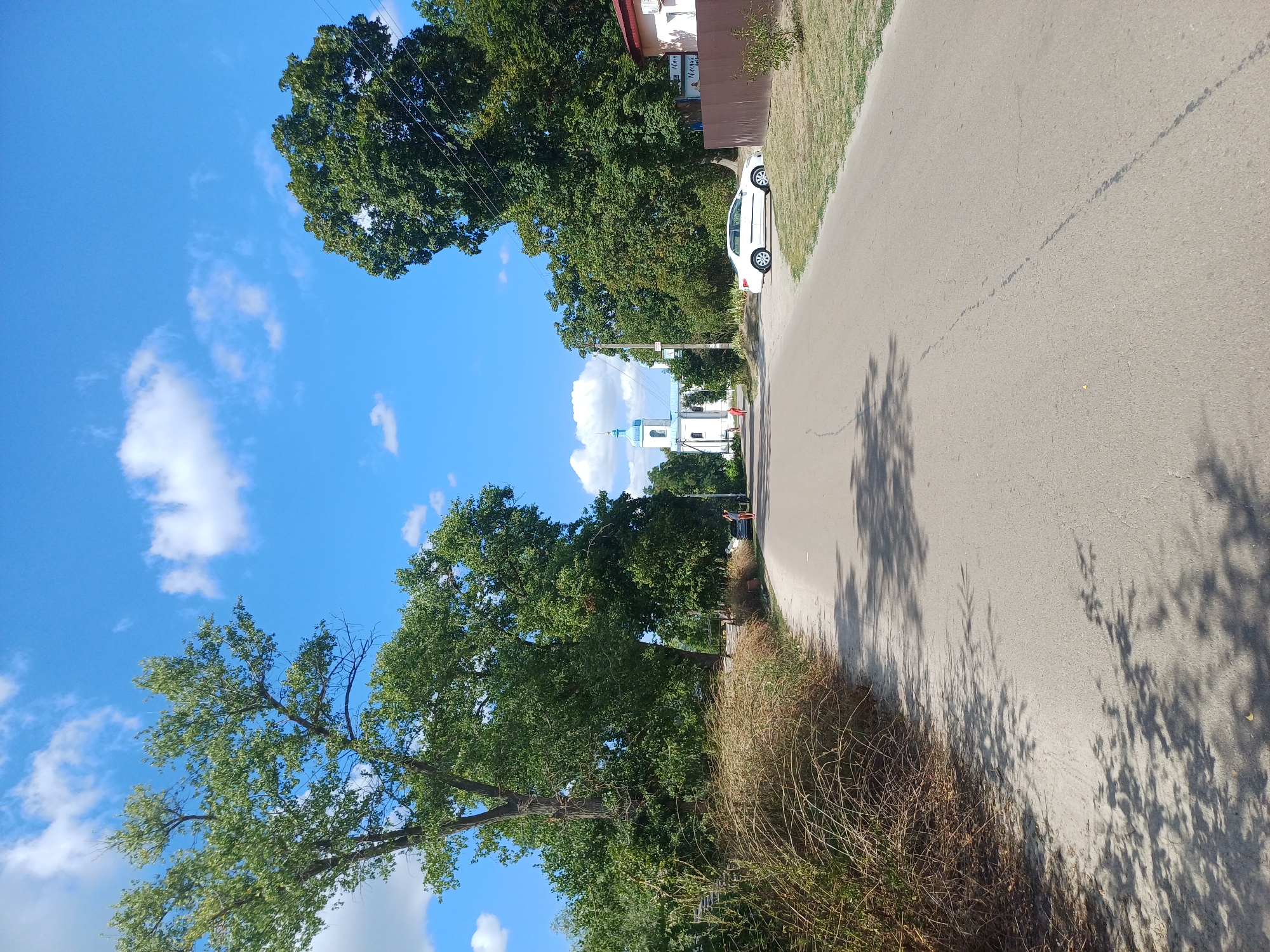 The image depicts a street scene in Sulymivka, Ukraine, on a bright, sunny day with a blue sky dotted with white clouds. A paved road runs towards a white church with a blue steeple in the distance. Lush green trees line both sides of the road. In the foreground on the left, there is dry, tall grass. On the right side of the road, a white car is parked. Two figures are visible walking in the distance on the left side of the road, and another figure is standing near the parked car on the right. The environment appears to be a rural or semi-rural area.