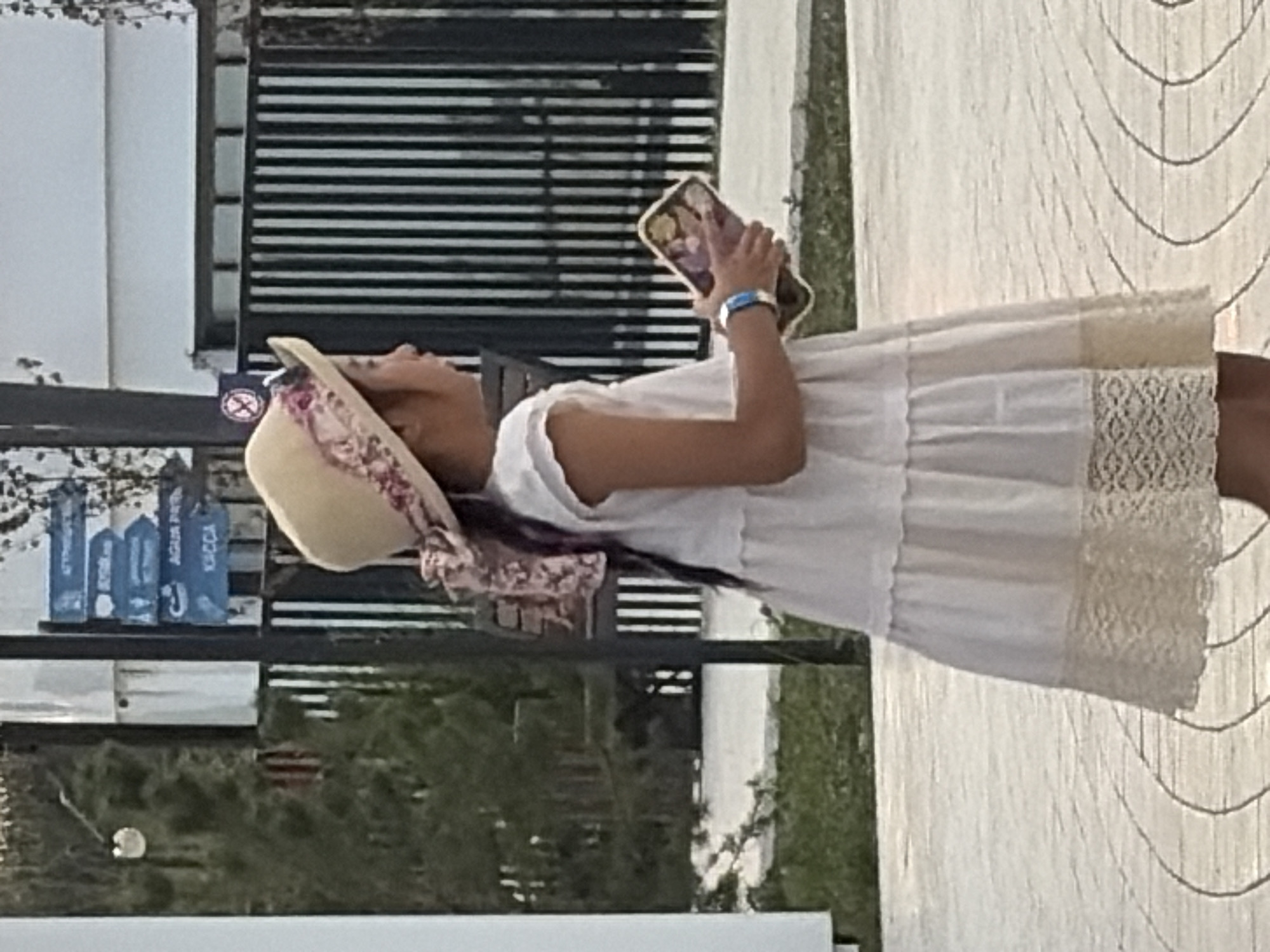 A young girl, facing right in profile, walks on a patterned walkway. She wears a white, tiered dress with a lace hem and a straw hat adorned with a floral ribbon. In her hands, she holds a mobile device with a colorful case. To her left, a dark pole with directional signs is visible, indicating "AGUA PARK" and "KACCA". Behind her, a black metal fence lines the background, with a white structure beyond it. The scene is outdoors, with a hint of greenery and trees on the left side of the image.