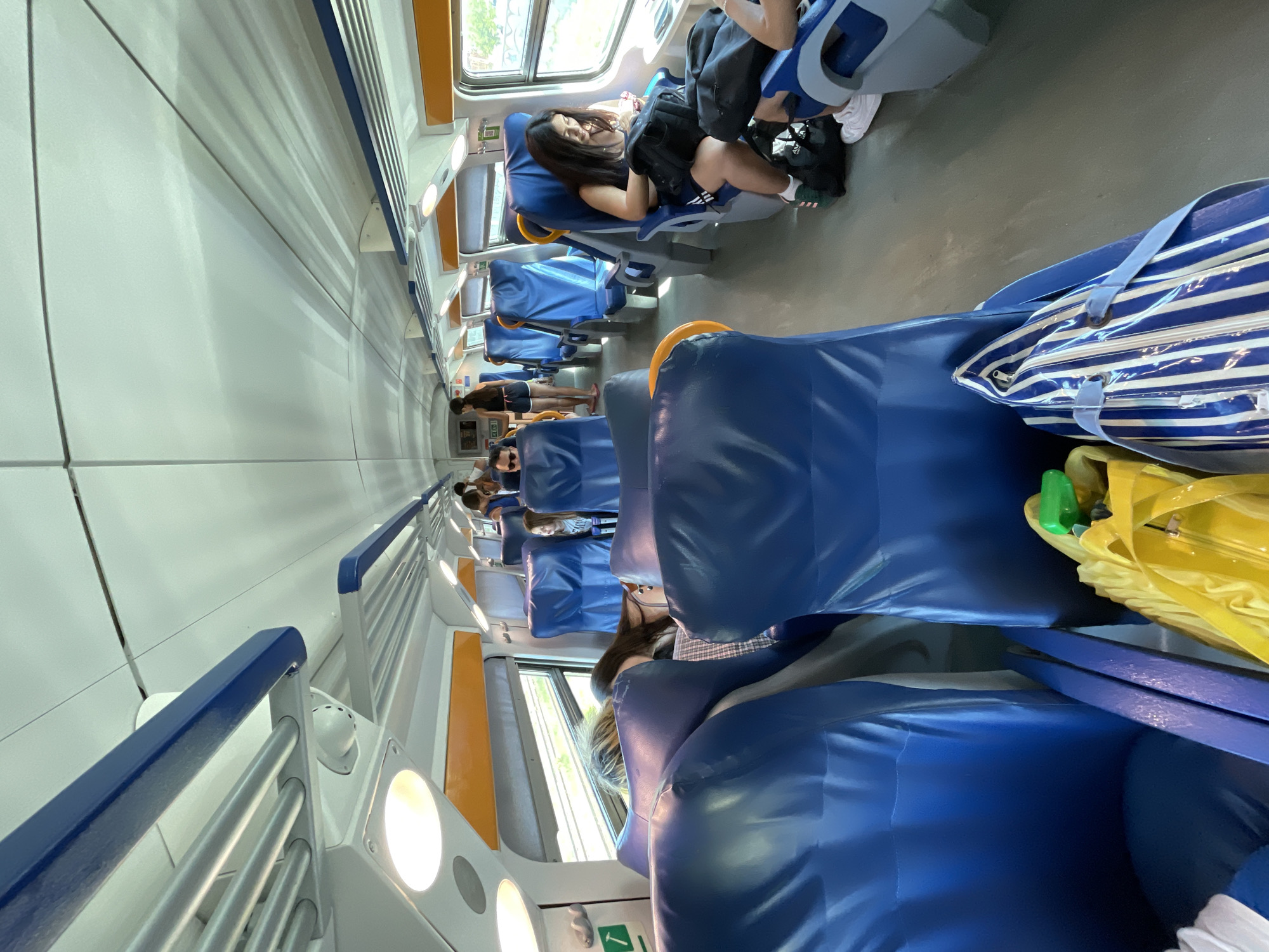 The image shows the interior of a train with rows of blue upholstered seats. Several people are visible, seated and standing. In the foreground, a yellow bag and a blue and white striped bag are on the floor next to the seats. On the right, a young woman is seated, looking at her phone. Further down the aisle, other passengers are seated. Overhead compartments and lighting fixtures are visible along the ceiling. The overall impression is of a typical train carriage during travel. Without additional information or context, it is not possible to definitively place this scene in Viareggio, Italy.