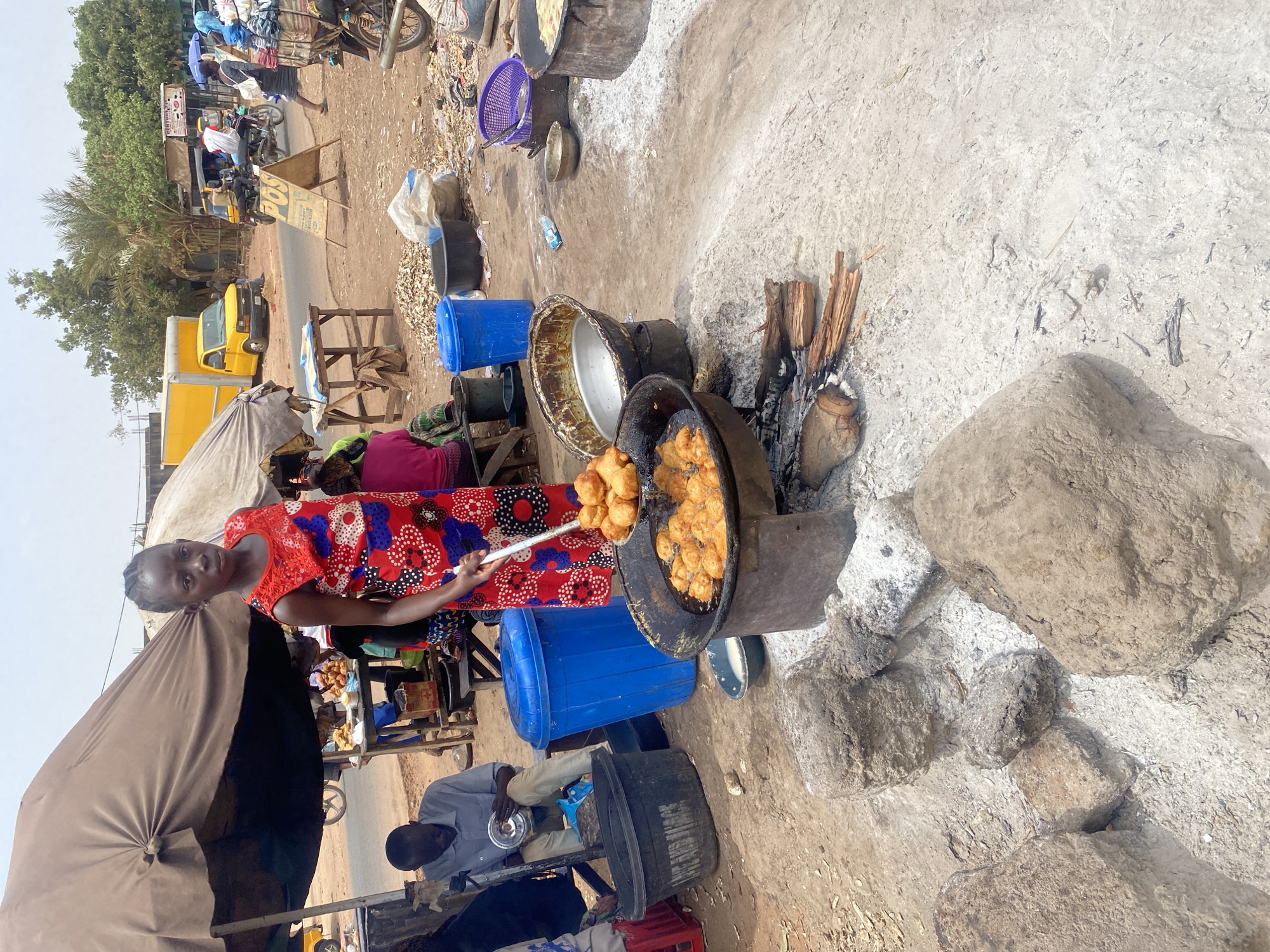An outdoor market scene in Rido, Nigeria, depicts a young female engaged in street food preparation. A dark-skinned female, approximately 10-14 years old, with braided hair and wearing a red, blue, and white patterned sleeveless dress, stands centrally. She holds a metal slotted spoon, lifting multiple golden-brown, spherical fried items (possibly puff-puff) from a large, black metal pot filled with hot oil. The pot rests on a makeshift stove composed of rocks and metal, fueled by burning wood logs at its base, surrounded by a grey ash residue on the dirt ground.
To the left, a man in a light blue shirt and tan trousers sits on a low wooden bench under a large, dark brown fabric umbrella, observing a metal dish. Nearby are two blue plastic cylindrical bins and a black plastic tub with faded text. Further into the background, beneath the umbrella, a wooden table displays various food items.
The midground features additional cooking stations, including another large pot over a fire, and several smaller metal and plastic bowls. The ground is dirt, covered in scattered debris including wood shavings, plastic bottles, and bags.
In the background, a paved road stretches diagonally, populated by a yellow delivery truck parked on the left, and several motorcycles (okadas) carrying riders and passengers. Roadside structures include a "POS" sign, rudimentary shops, and sparse vegetation under a hazy sky.