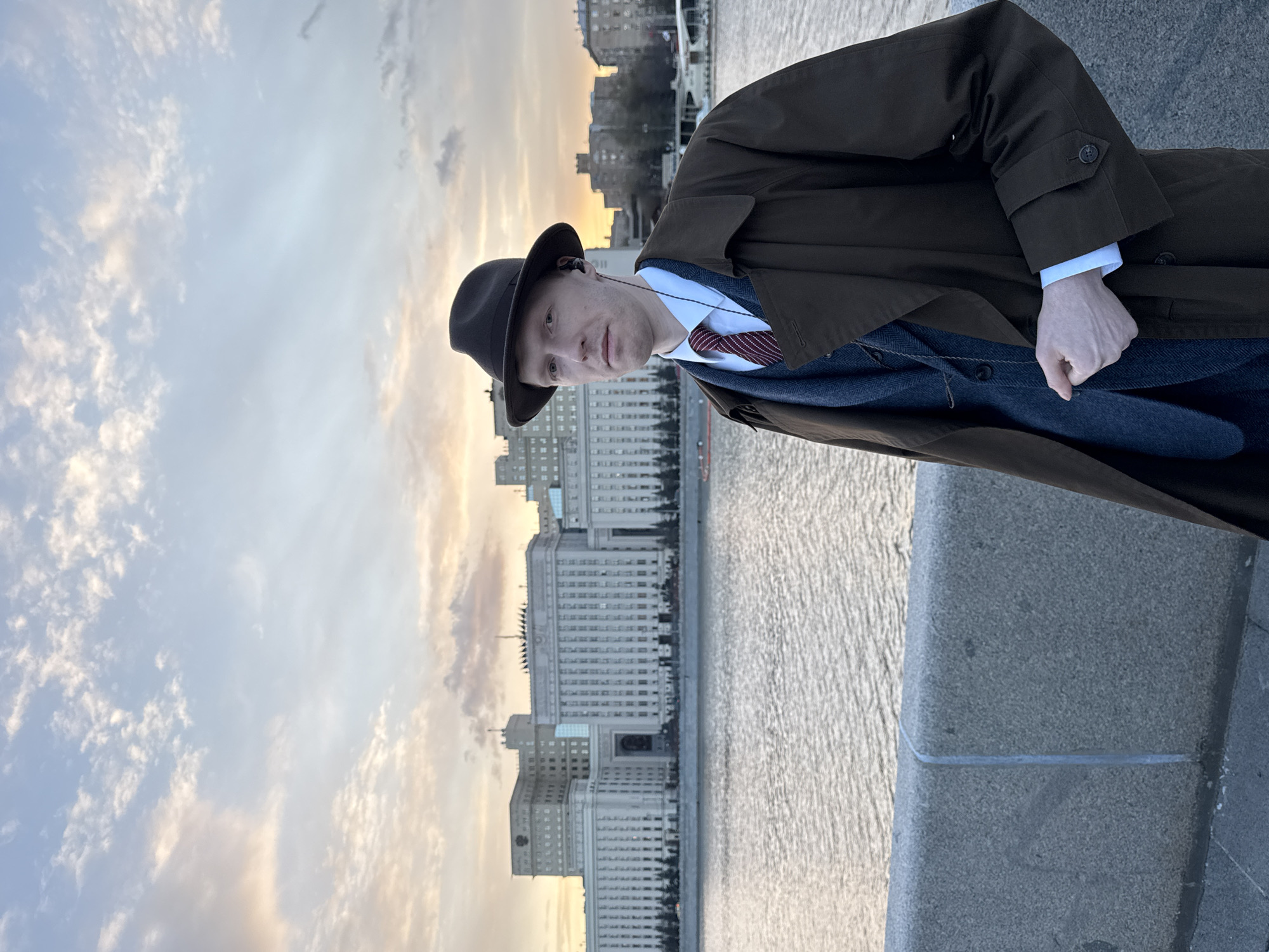 A man wearing a dark brown overcoat and fedora stands next to a river in Moscow, Russia. He has a pale complexion and wears a white collared shirt with a dark tie, a navy blue vest, and a suit jacket beneath his coat. In the background across the river is the Ministry of Defence building. The sky is filled with clouds and the setting sun casts a soft light. He is likely standing on a stone embankment.