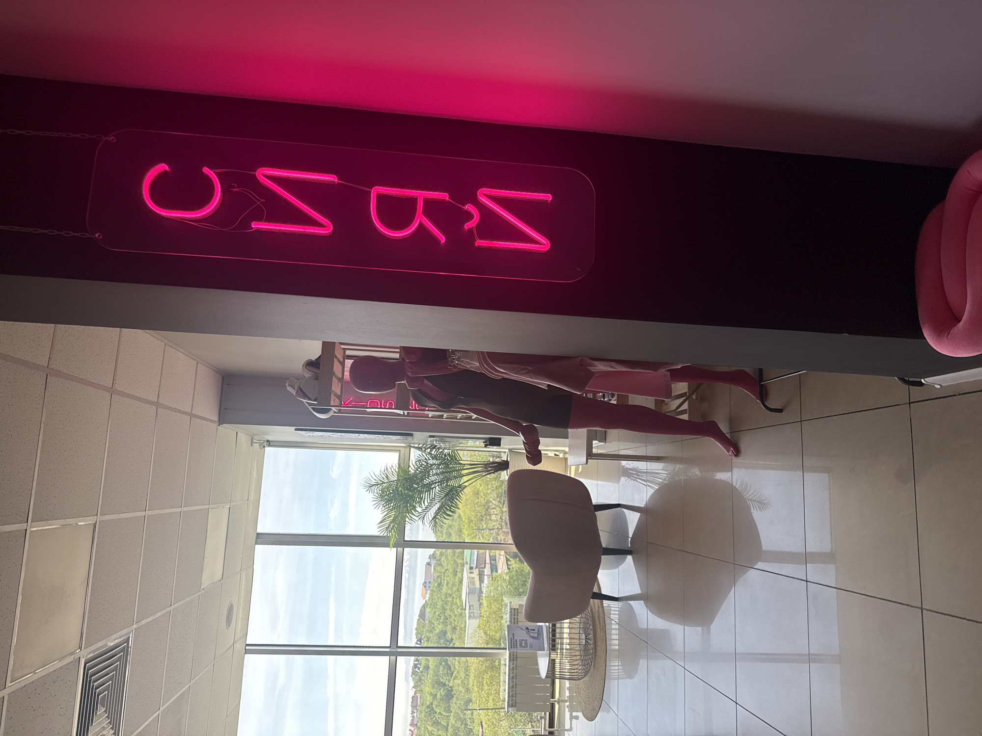 In Petropavlovsk-Kamchatsky, Russia, this image captures the interior of a retail space, likely a clothing boutique. A bright pink neon sign, spelling "СИЯЙ" (Shine) in Cyrillic, hangs vertically on a dark wall to the right. In the foreground, two mannequins are visible; one is a full-body figure wearing a dark dress and pink leggings, and the other is a partial view of a torso wearing a pink garment. The background reveals a spacious area with large windows overlooking a lush, green cityscape with buildings and hills under a cloudy sky. Inside, a modern pink armchair sits next to a circular coffee table with a white tabletop, all on a highly reflective tiled floor. A potted palm adds a touch of greenery to the scene. The overall impression is a stylish and contemporary retail environment.