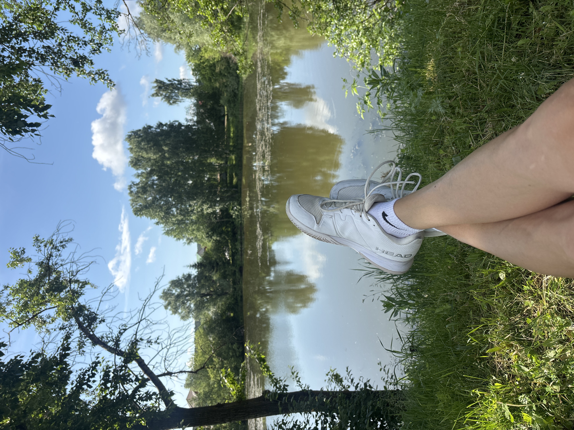 A person is reclining on a grassy bank next to a body of water, likely a lake or river, on a sunny day. The person's legs are crossed at the ankles, with white tennis shoes prominently displayed. The shoes have the brand name "HEAD" visible on the side. The background consists of trees lining the opposite bank of the water, with their reflections visible in the water. The sky is blue with scattered clouds. Based on the vegetation and overall environment, the location is likely a park or recreational area near water. It is impossible to determine if the location is in the city of Chaikovsky, Russia based on visual information.