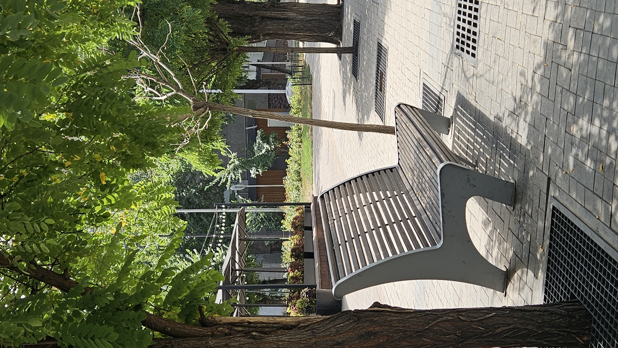 The image depicts a park bench situated on a paved walkway in Almaty, Kazakhstan. The bench is made of wood slats and has a gray metal frame. It is positioned under a canopy of green trees, casting dappled shadows on the ground. The walkway is made of light-colored paving stones, and several metal grates are visible, likely for drainage. In the background, there are more trees, some buildings, and a covered outdoor seating area with umbrellas. The scene appears to be a public space designed for relaxation and leisure. There are no people present in the image.