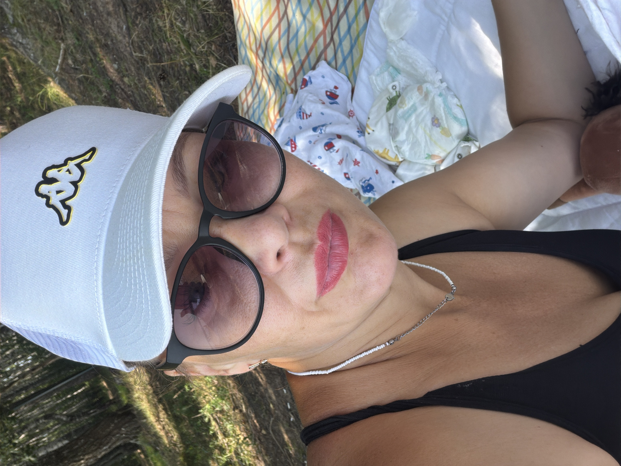 In a close-up, over-the-shoulder selfie, a woman with fair skin, rosy lips, and dark-colored sunglasses is positioned in the foreground. She is wearing a white baseball cap with a black and gold logo, a black tank top, and a beaded necklace. Her face is partially obscured by her sunglasses, but her mouth is slightly open. The background shows a natural setting with trees and foliage, suggesting an outdoor location. A striped blanket and some baby items, including a diaper, are visible to her right. The image appears to be taken during daylight. The mention of Chaikovsky, Russia is not supported by visual evidence in the image.