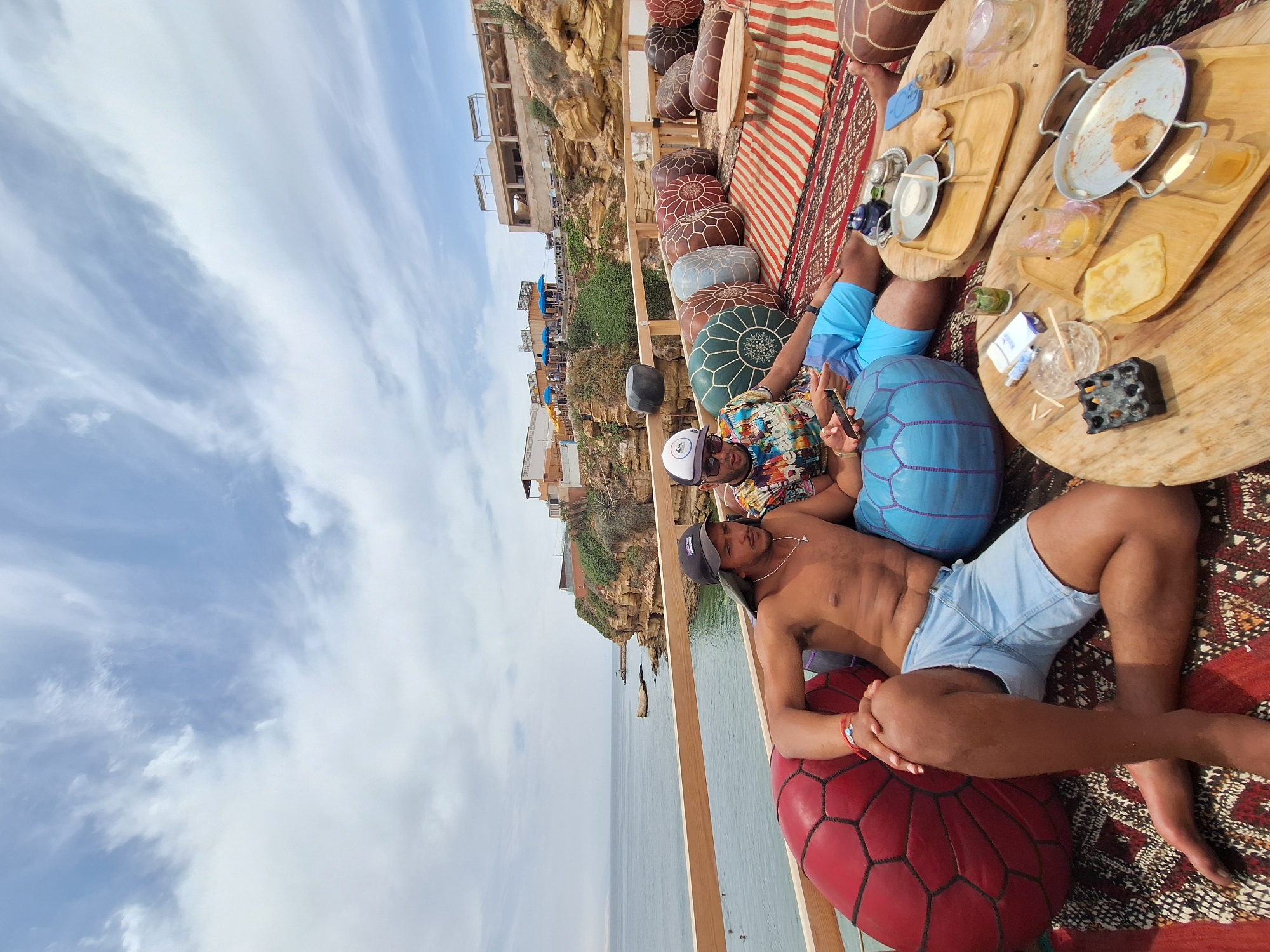 Two men are seated on large floor cushions with a rug and a wooden table in front of them. The man on the left is shirtless, wearing light blue shorts and a black baseball cap. The man on the right is wearing a colorful patterned shirt and a white baseball cap. They are on an outdoor terrace overlooking the ocean with a rocky cliff face to the right. Buildings are visible on the cliff. The table holds plates with food, glasses, and a teapot. The setting appears to be a cafe or restaurant in Imsouane, Morocco.