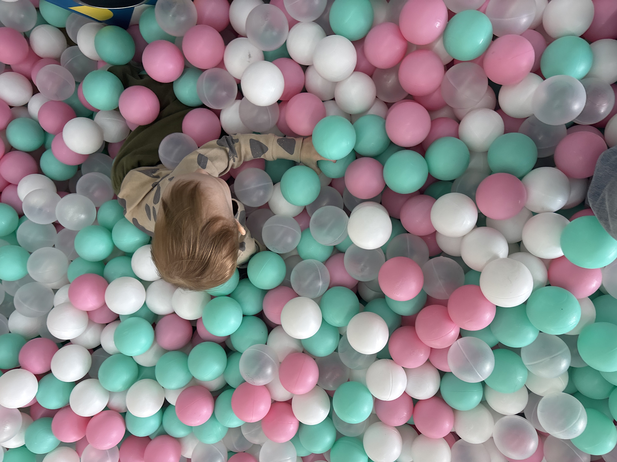 A top-down shot shows a toddler partially submerged in a ball pit. The ball pit contains white, pink, and teal-colored balls. The toddler has short blond hair and is wearing a tan shirt with a camouflage pattern. The toddler's face is obscured, but the back of their head and one arm are visible. The location is in the city of Perm, Russia.
