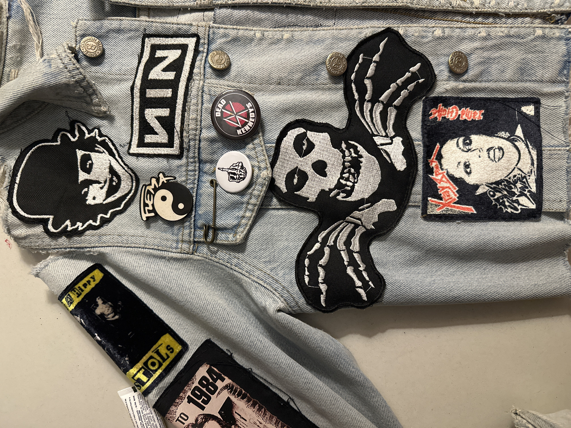 The image shows a close-up of a light blue denim jacket adorned with various patches and pins. Patches include NIN (Nine Inch Nails), a cartoon head, a skull with skeletal wings (likely a Misfits patch), and a patch with Japanese text and a graphic. Pins feature bands Dead Kennedys and an image of a raised middle finger. Additional patches on the lower portion include a Sex Pistols patch and one featuring the text "1984." The garment appears to be lying flat on a surface. There are no indications of the location in Buenos Aires, Argentina.