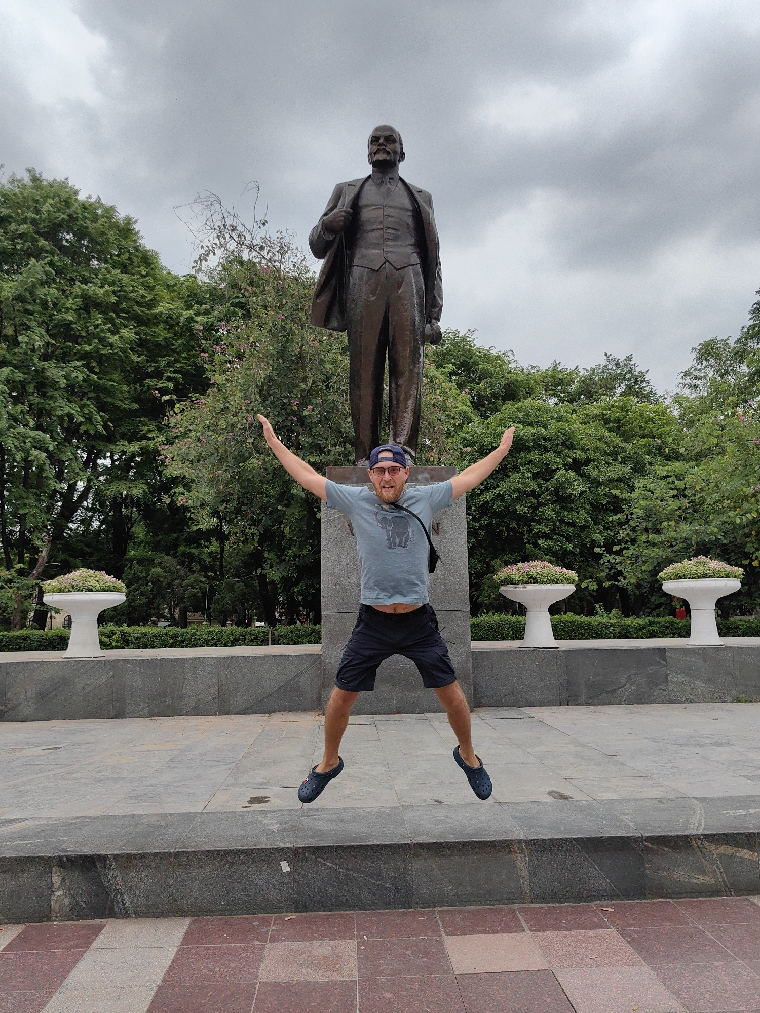 The image displays an outdoor scene in Hà Nội, Vietnam, featuring a man jumping playfully in front of a prominent historical statue. A fair-skinned man, wearing a blue t-shirt with an elephant graphic, dark shorts, blue Crocs, a blue baseball cap, and glasses, is captured mid-air with his arms spread wide and legs bent. He has a beard and mustache and appears to be smiling or laughing, conveying a cheerful and energetic emotion.
Behind the man stands a large bronze statue of a figure resembling Vladimir Lenin, depicted in a suit and vest, positioned on a tiered stone pedestal. The setting is a public park or plaza, characterized by lush green trees and bushes forming the background. Two ornate white planters, filled with small green and pink flowering plants, are placed on either side of the statue's base. The ground is paved with a combination of grey stone and reddish-brown and beige checkered tiles. The sky is overcast, suggesting cloudy weather and providing soft, diffused daylight. There is no visible text in the image.