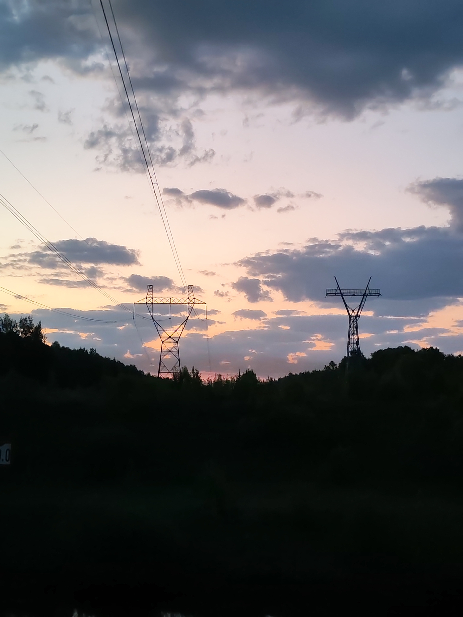 The image depicts two electrical transmission towers silhouetted against a sunset sky with scattered clouds. The towers are situated behind a dense line of trees, suggesting a rural or suburban location. Electrical wires are strung above the leftmost tower. The foreground is dark, obscuring details. The location is assumed to be near Zaprudnya, Russia, based on the prompt, but the image itself contains no specific indicators of location.