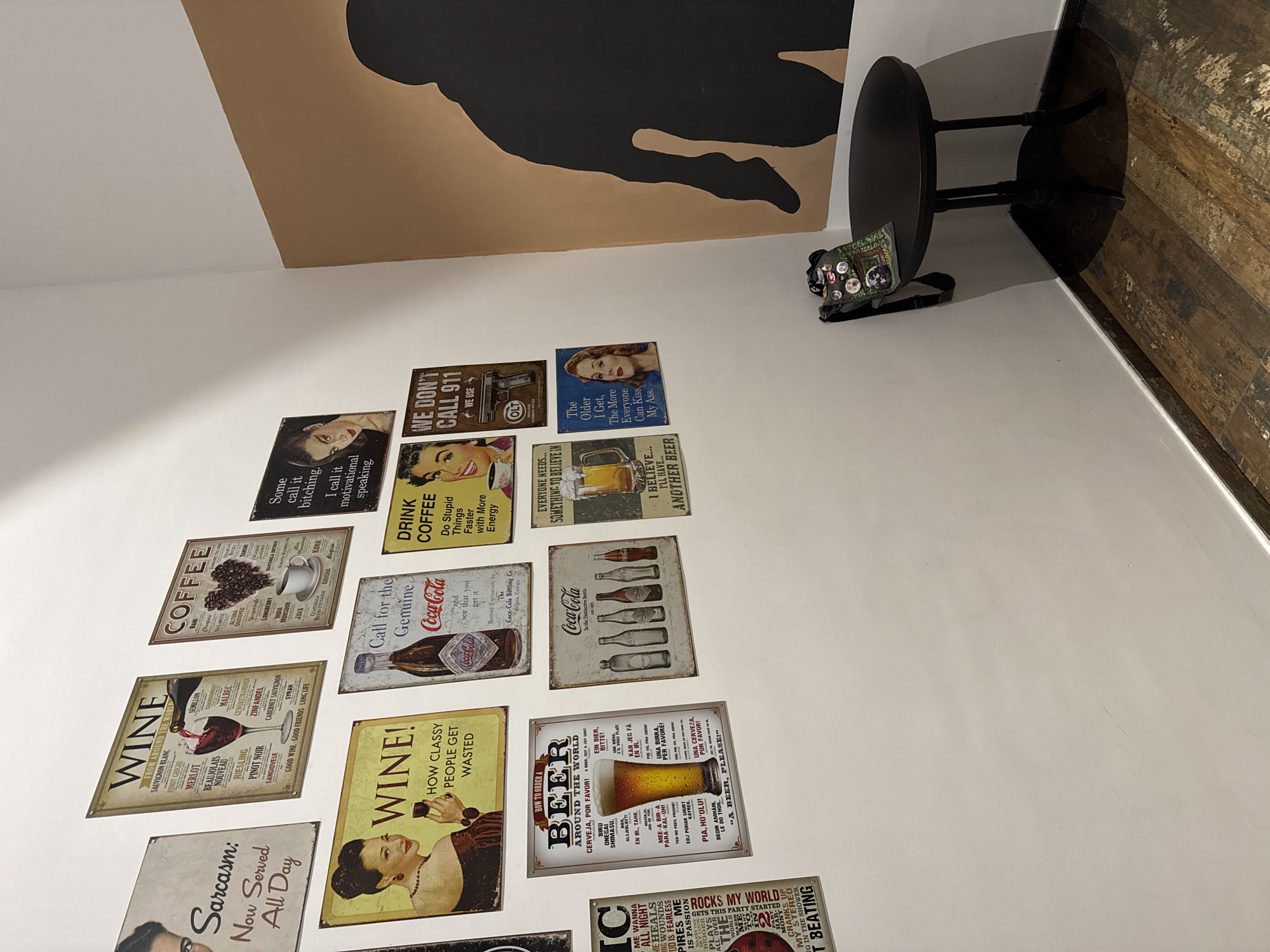 A photograph taken inside a bar or restaurant, showing a white wall covered in vintage-style beer and wine posters. A small, black, round table stands in the corner near the wall, with a small black bag with patches resting on the table. An abstract painting with a figure is mounted above the wall decor. The floors are partially visible to the right and appear to be made of wood.