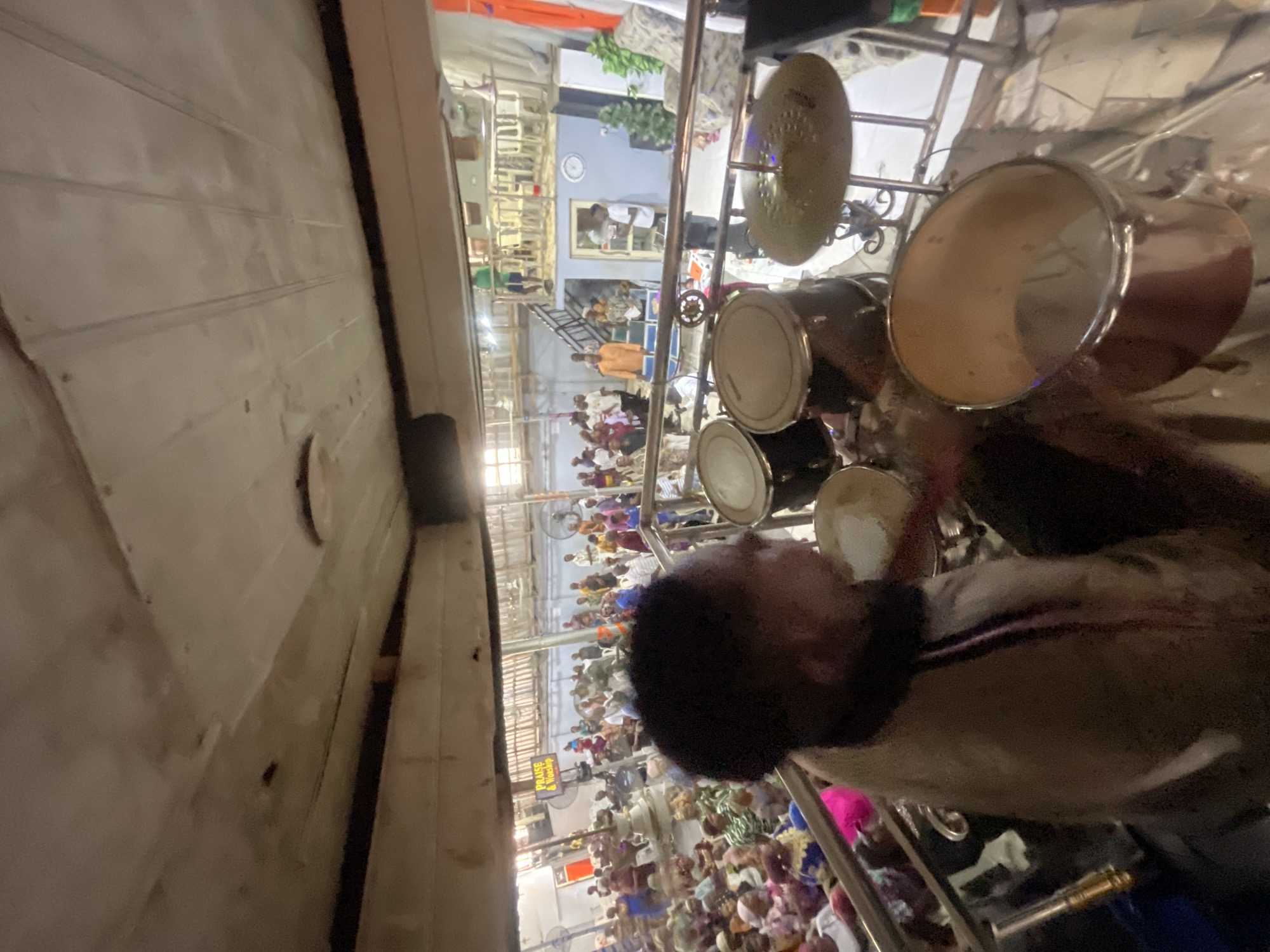 In Alagba, Nigeria, a drummer is playing a drum set inside a building, possibly a church or community hall, during a service or event. The drummer, a person of color, is positioned in the foreground, with their face partially obscured by the low-angle perspective. They are wearing a light-colored jacket with darker trim. The drum set includes at least two toms and a cymbal, with part of a large floor tom visible in the lower right corner.
In the background, a large congregation of people are seated, facing forward, suggesting a religious gathering or performance. The architecture of the building is visible, with exposed beams and a ceiling that appears to be made of wooden planks and some plaster or concrete. There is a raised platform or balcony in the background with chairs set up, and several people are standing on it. One individual in a green shirt is visible on this elevated area. Further back, fans are mounted on the wall, indicating a warm climate. Signs in the background are partially visible, with one reading "PRAISE & Worship." The overall lighting suggests an indoor setting with artificial lights.