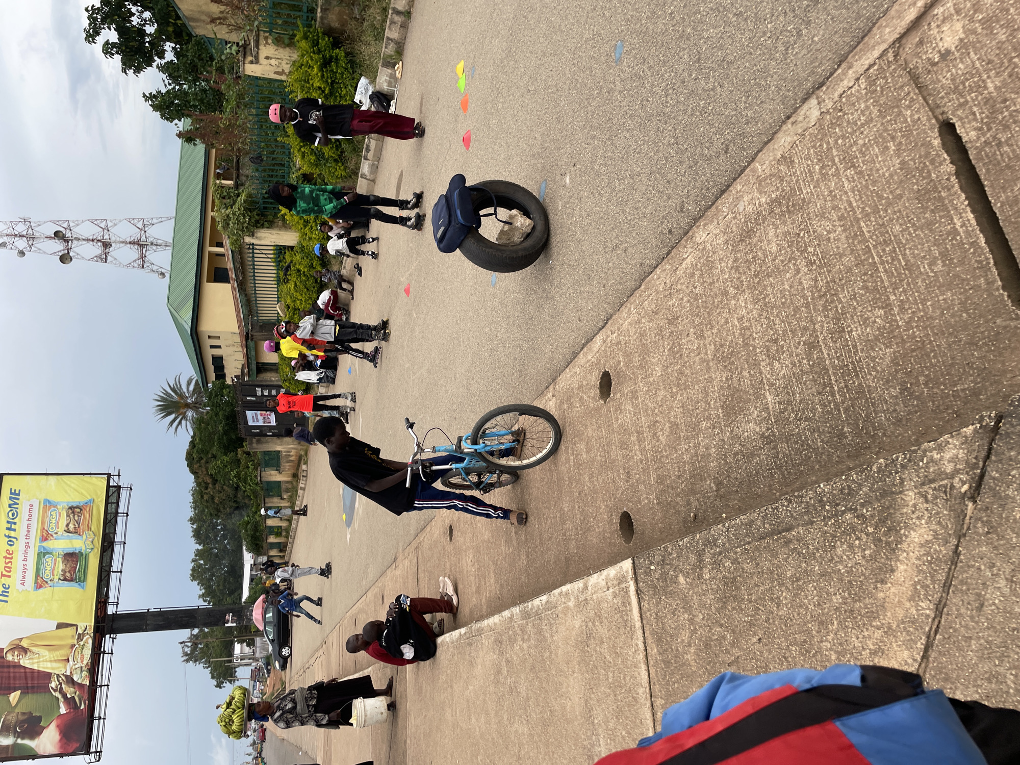 The image depicts a street scene in Jos, Nigeria, on an overcast day. In the foreground, a person in a blue and red jacket is partially visible, facing away from the camera. A light blue bicycle is parked on the concrete sidewalk next to a person wearing a black t-shirt and dark pants, who is bent over the handlebars. Further down the sidewalk, two young individuals are seated.
On the street, several children and young adults are engaged in activities. Some are wearing rollerblades, and one person appears to be wearing roller skates. A bright orange t-shirt stands out amongst the other clothing. In the background, a billboard advertises "The Taste of HOME" with images of food products. A cellular tower is visible in the distance, as are buildings with green roofs and fences. The street is lined with vegetation and trees. Several colorful cones are scattered on the asphalt, possibly marking a course for the rollerblading activities. A tire with a backpack and a rock inside is placed in the middle of the road. A woman carrying a large bunch of bananas on her head walks along the sidewalk carrying a white bucket.