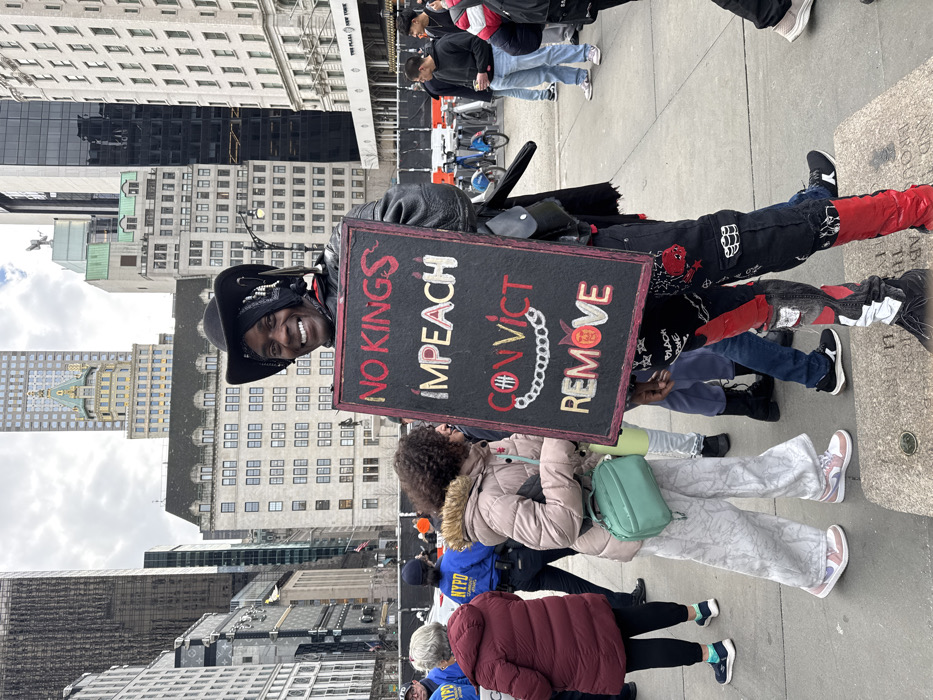 A smiling man in a cowboy hat holds a sign stating "NO KINGS IMPEACH CONVICT REMOVE" with a drawn chain connecting "CONVICT" to "REMOVE." He stands in front of a crowd on a city street with tall buildings in the background. The man is wearing black and red pants and a black jacket. He is the focal point of the photo.