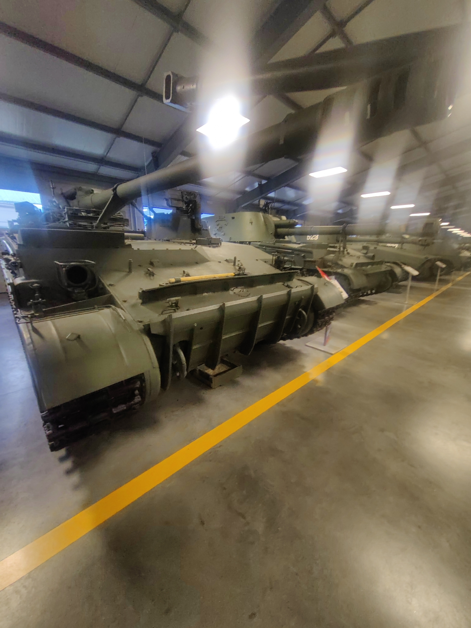 The image shows the interior of a museum or exhibition hall, likely the Patriot Park in Kubinka, Russia, which houses a large collection of military hardware. In the foreground, the left side of a green military tank is visible, facing towards the right of the frame. The tank has a long barrel, a turret with some visible details, and a robust tracked undercarriage. The hull features various hatches, panels, and what appears to be a towing pintle at the front. A bright yellow line runs diagonally across the concrete floor, separating the display area from a walkway. In the background, the blurred shapes of other military vehicles, including at least one other tank and possibly an armored personnel carrier, are arranged in a line. The environment is illuminated by overhead artificial lighting, and the ceiling structure, with exposed beams and panels, is visible. The overall impression is one of a carefully curated exhibition of heavy military equipment within a large, industrial-style building.