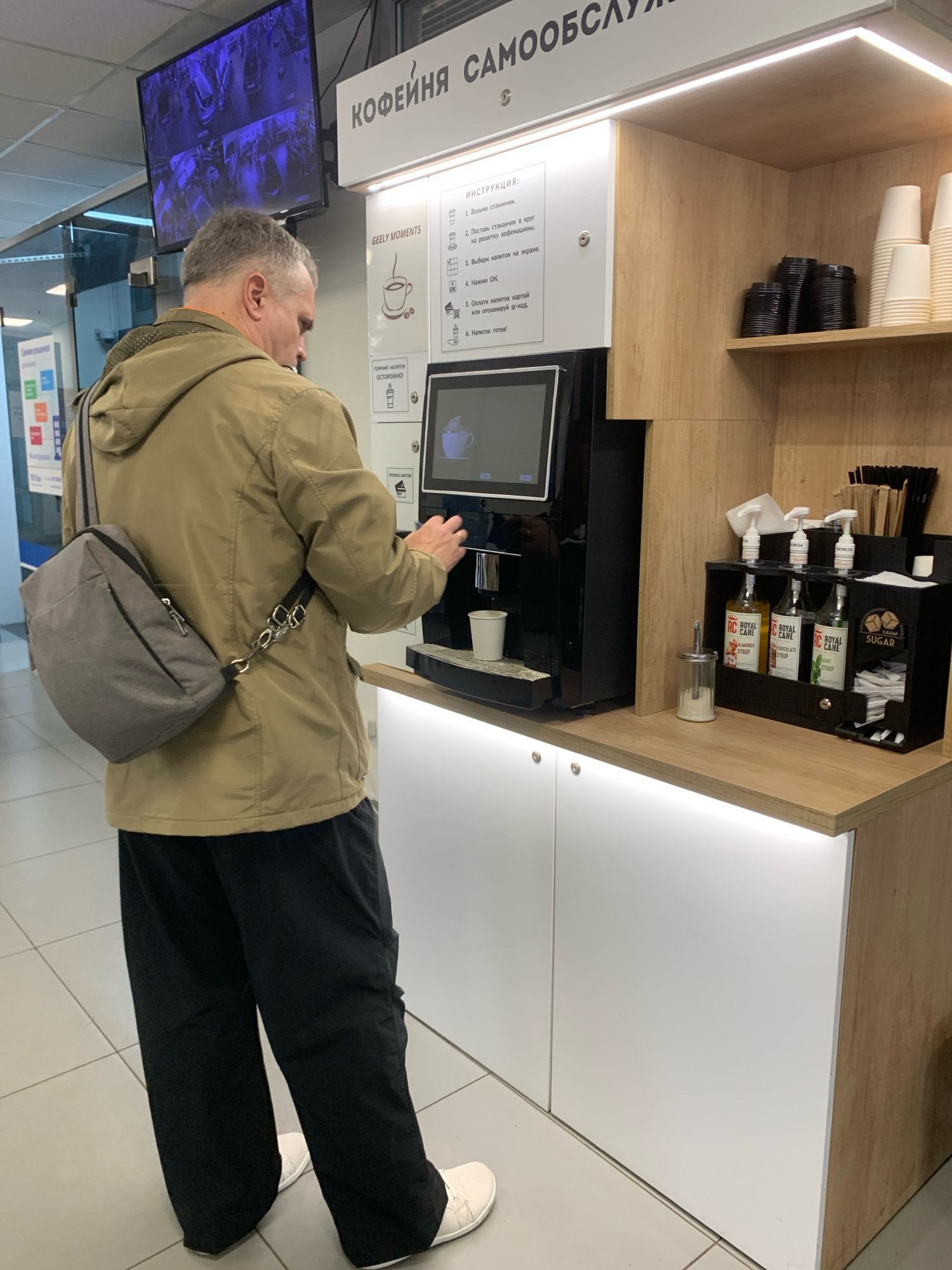 A middle-aged man with gray hair is standing in front of a self-service coffee machine. He is wearing a light brown jacket, black pants, and white shoes. He has a gray crossbody bag over his left shoulder. The man is reaching out towards the coffee machine, possibly to select a drink or add something to his cup.
The coffee machine is black with a touchscreen interface. Next to the machine, there are shelves stocked with paper cups, coffee pods, and various syrups in bottles labeled "ROYAL CANE" with flavors like "ALMONDS SYRUP," "CHOCOLATE SYRUP," and "MINT SYRUP." There is also a dispenser of sugar packets and a sugar shaker. A sign above the machine has instructions in Russian. A TV screen displaying security camera footage is mounted on the wall above the coffee station.
The environment appears to be an indoor public space, possibly an airport or a train station, given the signage and the overall setup. The lighting is bright, and the floor is tiled. The overall impression is one of a modern, functional self-service area. The location is the city of Minsk, Belarus.