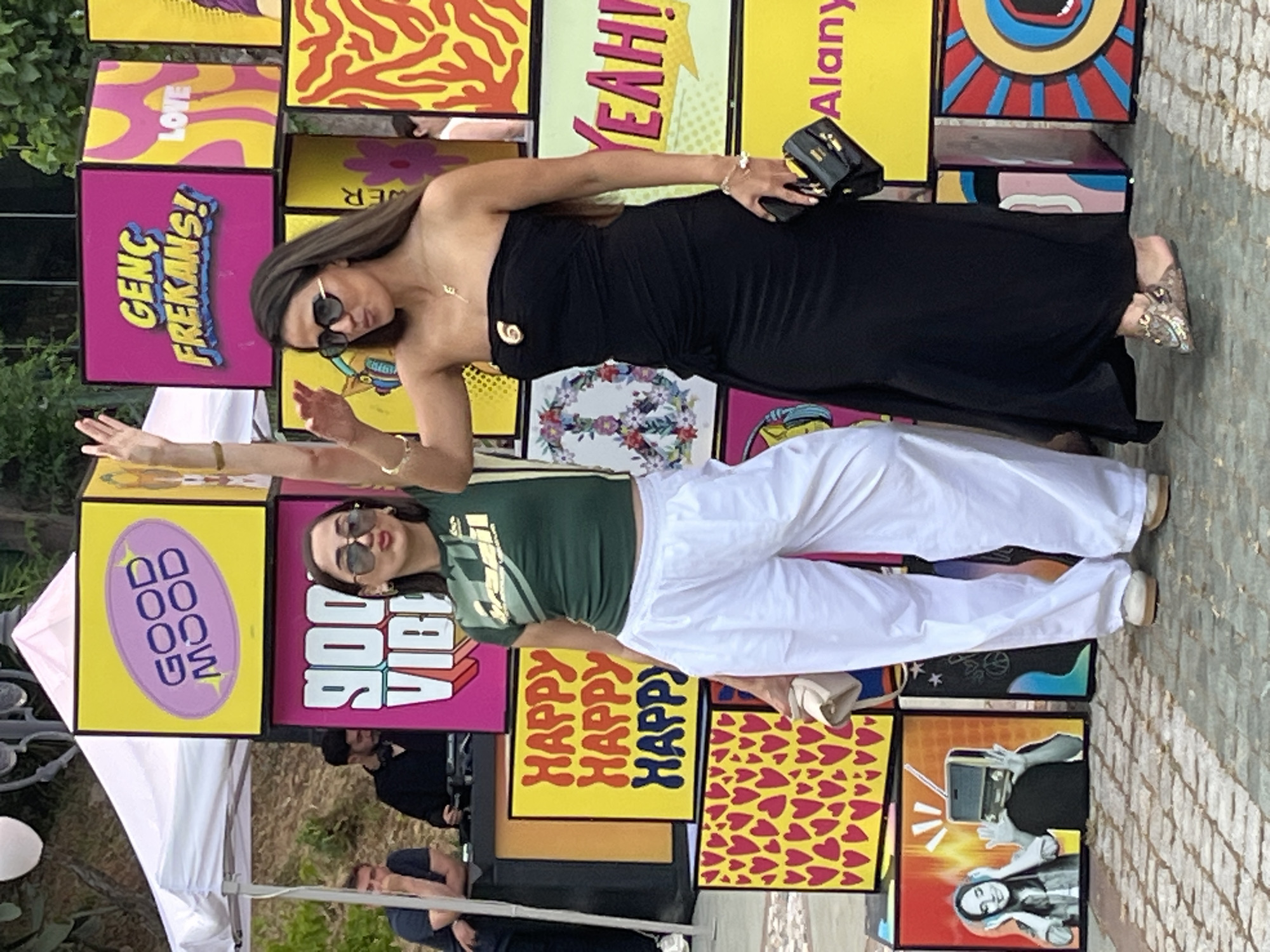 The image depicts two women posing in front of a wall constructed of colorful, pop-art-inspired boxes. The woman on top wears a strapless black dress and holds a small black purse. She also wears sunglasses and a necklace. The woman below wears a green t-shirt with gold accents and white pants. She also wears sunglasses and a bracelet. The boxes behind them have words and phrases like "GOOD MOOD," "900 VIBE," "GENC FREKANS!" "LOVE," "YEAH!" and "HAPPY." One box reads "Alany," suggesting this is a location within the city of Alanya, Turkey. Part of a white tent is visible on the left. Other people are in the background near the tent. The ground appears to be paved with cobblestones.