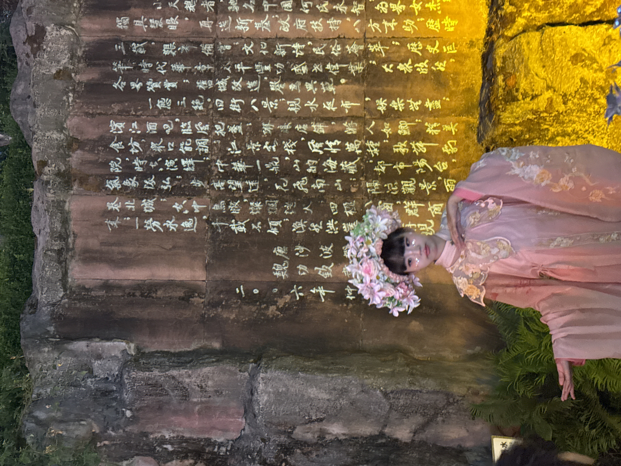 In the Yuzhong District of China, a young girl dressed in a pink, traditional Chinese outfit stands before a rock face inscribed with white calligraphy. She wears a floral headdress and has glitter on her face. The rock face is illuminated from below by a warm yellow light, creating a dramatic contrast with the dark greenery in the foreground. The calligraphy appears to be ancient text, adding a historical and cultural element to the scene. The overall impression is one of performance or cultural presentation in an outdoor setting.