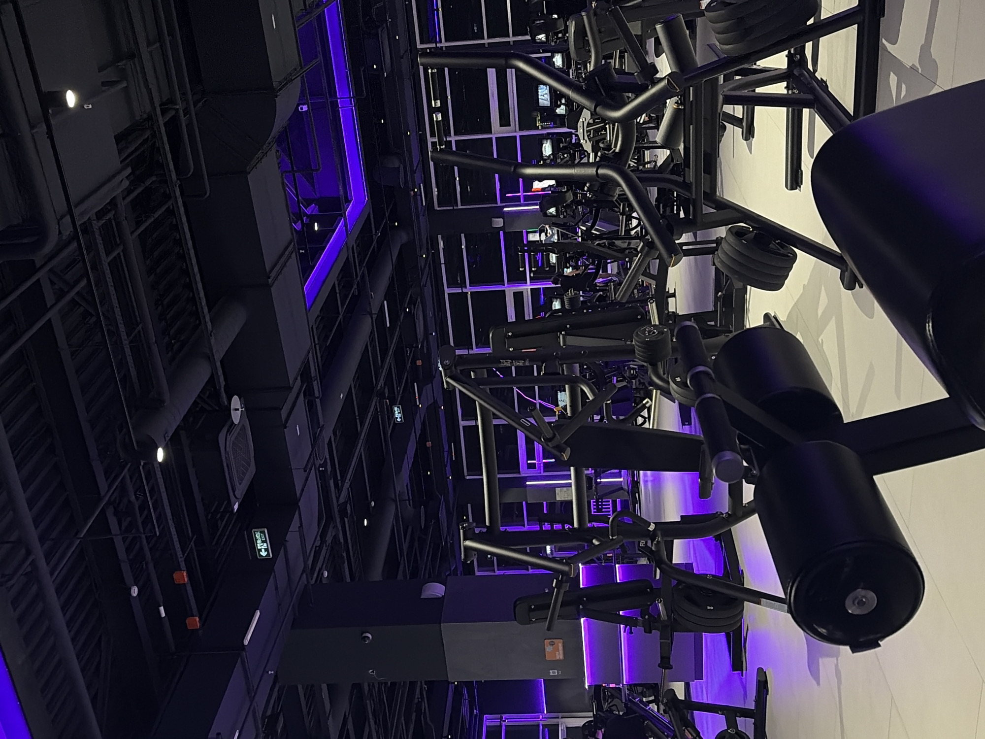 The image depicts the interior of a modern gym, characterized by its dark color scheme with prominent purple lighting accents. The gym is equipped with a variety of black exercise machines, including weight machines and what appear to be cardio machines in the background. The flooring is a light-colored tile or polished concrete, reflecting the ambient lighting. The ceiling features exposed ductwork and a complex lighting system, with some areas illuminated by vibrant purple LED strips. There are no discernible people in the foreground, but faint silhouettes of individuals can be seen near some of the machines in the distance. An illuminated green exit sign with Russian text ("ВЫХОД EXIT") is visible on the ceiling towards the left side of the image. The overall impression is one of a contemporary and well-lit fitness facility. The location is identified as Krasnoyarsk, Russia.