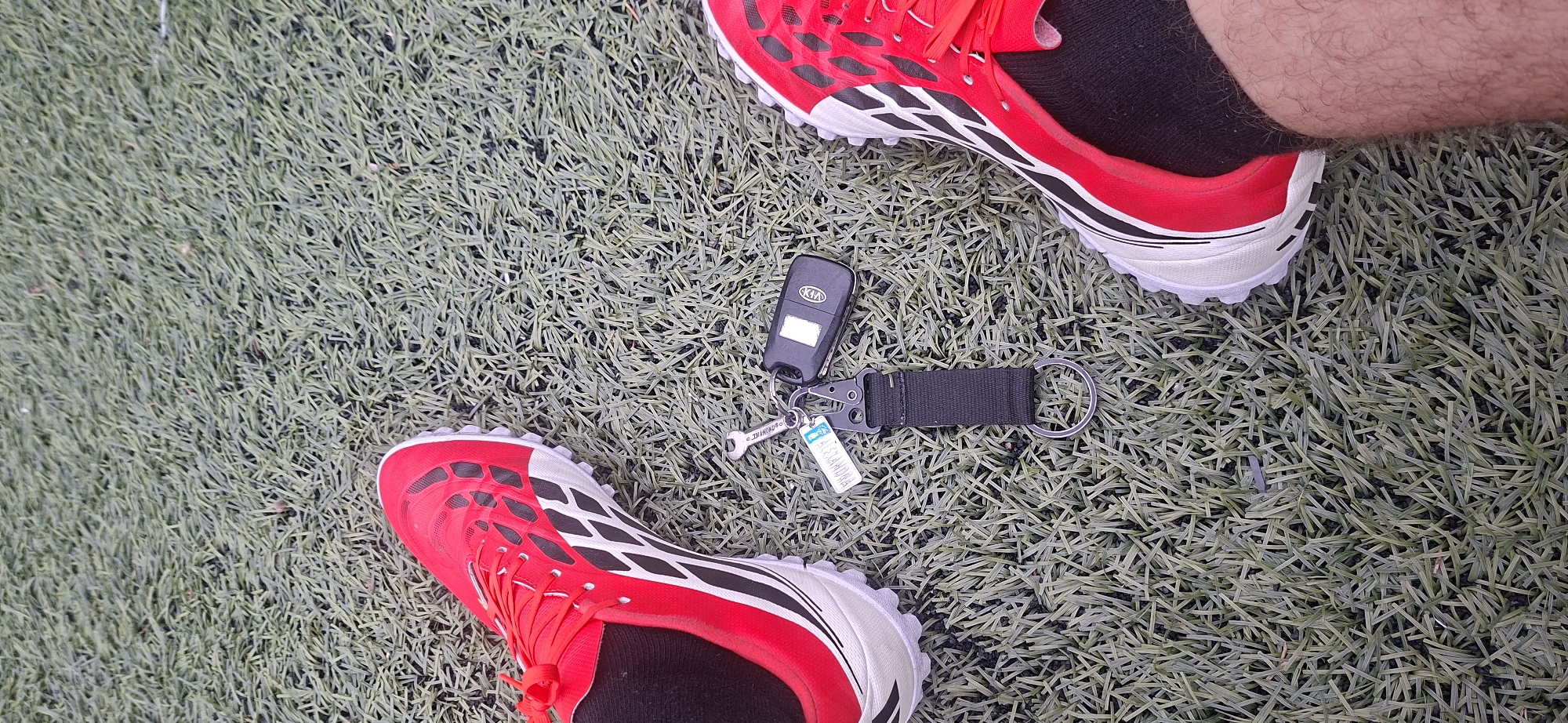 The image presents a top-down view of a person's feet and car keys resting on an artificial turf surface. The subject is a person, with portions of their lower legs and feet visible. They are wearing black socks and striking red and white athletic shoes, consistent with turf shoes or soccer cleats, suitable for artificial grass. The right leg shows some hair.
Positioned between the feet on the turf is a black Kia car key fob. It is attached to a keychain ensemble that includes a black fabric strap, a metal carabiner-style clip, and a large metal split ring. Also connected to the keychain is a small, silver-colored metal charm shaped like a wrench, bearing the inscription "DIAMOND®". A small blue and white tag is also part of the keychain, with the word "LUCKY" clearly visible at the top, followed by an unreadable alphanumeric string.
The setting is an outdoor artificial turf sports field or pitch, characterized by its uniform green synthetic grass blades. The lighting suggests it is daytime, casting a bright illumination without strong, defined shadows. The presence of athletic footwear on a sports field implies the person is engaged in, or pausing from, a sporting activity. The scene is set in Lyaskovets, Bulgaria.