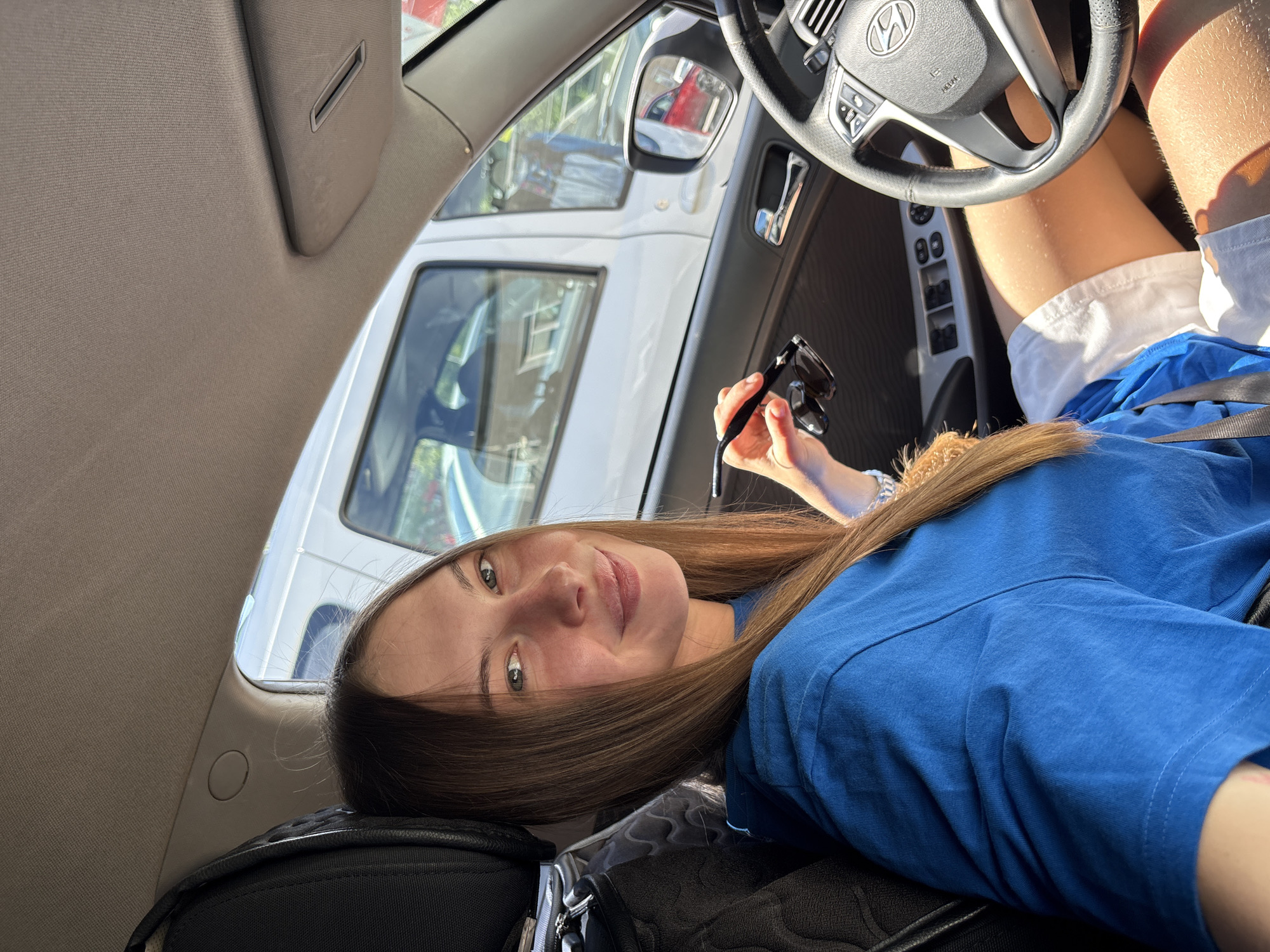 A young Caucasian woman with long, straight brown hair is seated in the driver's seat of a car. She is wearing a blue t-shirt and white shorts. Her left hand is holding a pair of dark sunglasses. She is smiling and looking at the camera. The interior of the car is visible, including the dashboard, steering wheel, and door panel. Through the windows, other vehicles and a building can be seen. The scene appears to be outdoors on a sunny day. The specific location within the city of Perm, Russia, cannot be determined from the image alone.