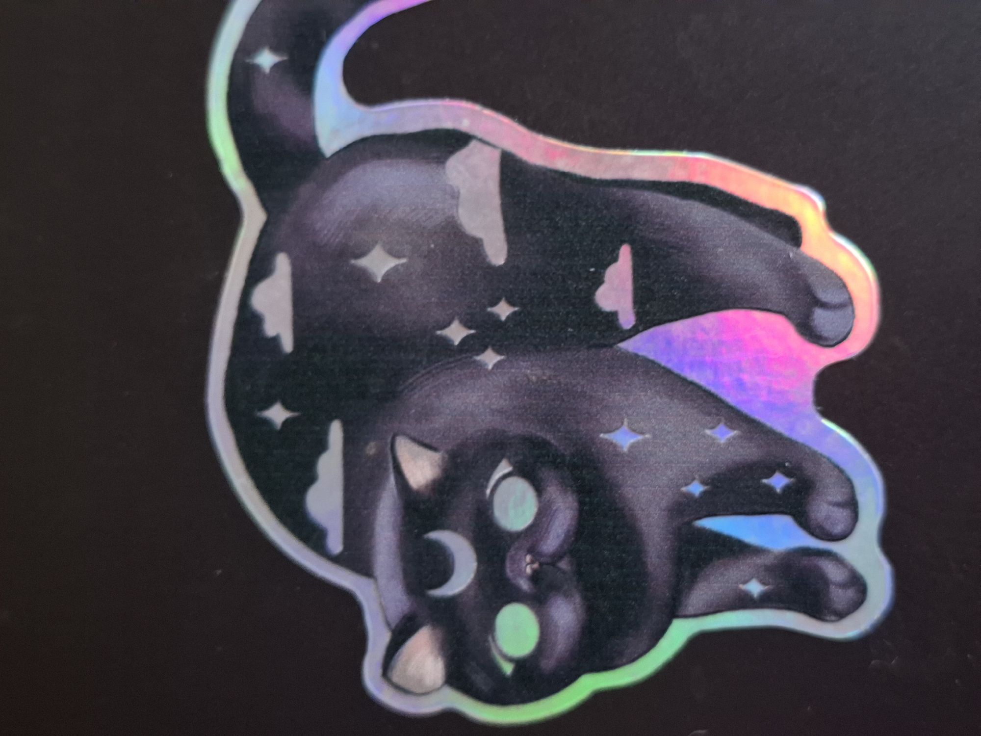 The image shows a holographic sticker of a black cat, oriented upside down, on a dark surface. The cat has a crescent moon marking on its forehead and is adorned with star and cloud shapes throughout its body. The edges of the sticker exhibit a rainbow holographic effect. There is no indication of the location being Novi Sad, Serbia.