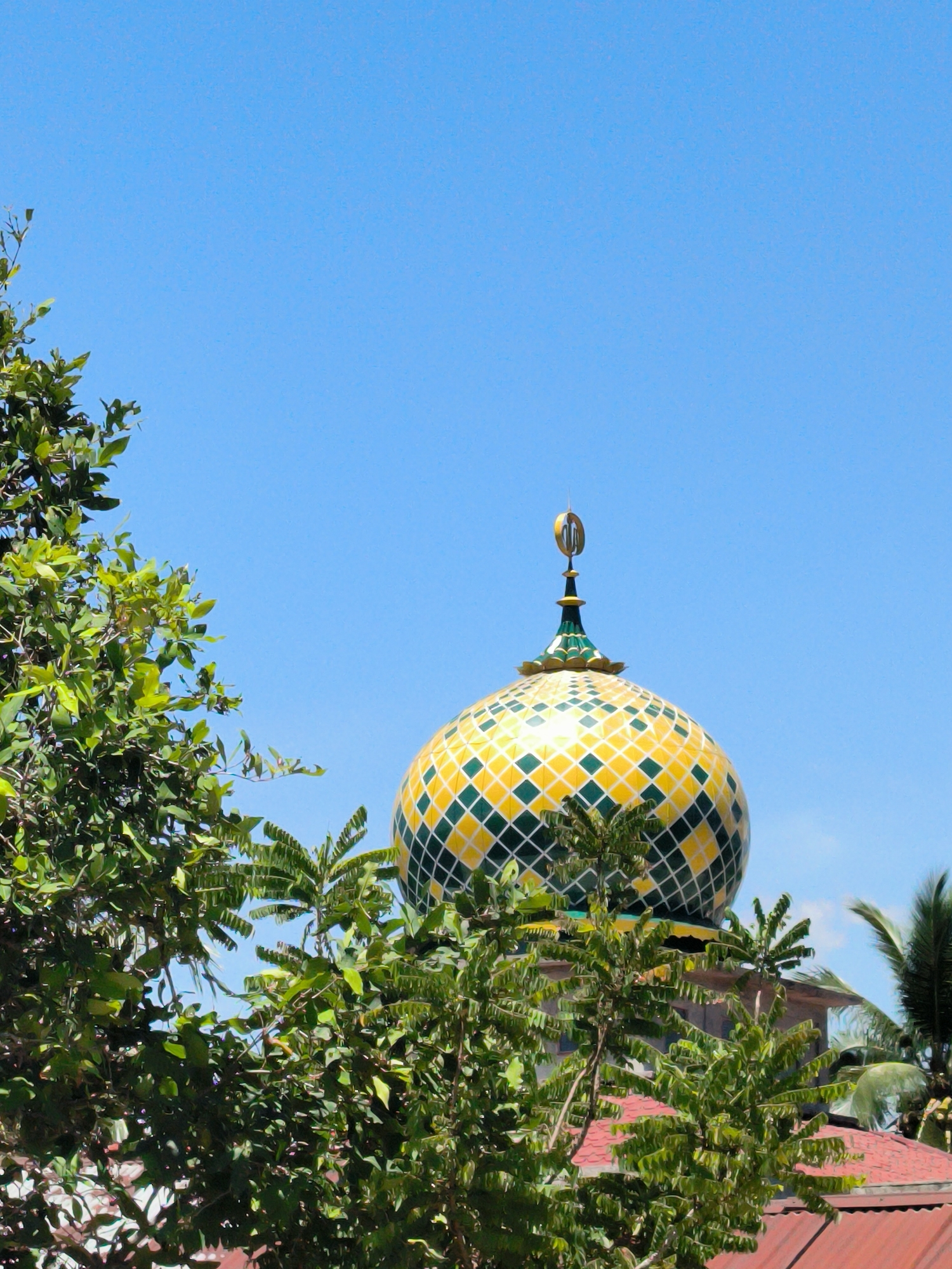 The photograph captures the top of a mosque dome against a clear blue sky. The dome is decorated with a pattern of yellow and green diamond shapes. At the very top, there is a golden crescent moon and star symbol. The dome is partially obscured by lush green foliage in the foreground, including tree branches and leaves. The background reveals a red tiled roof of an adjacent building, with palm fronds visible in the distance. The location of the image is Merbopuntong, Indonesia.