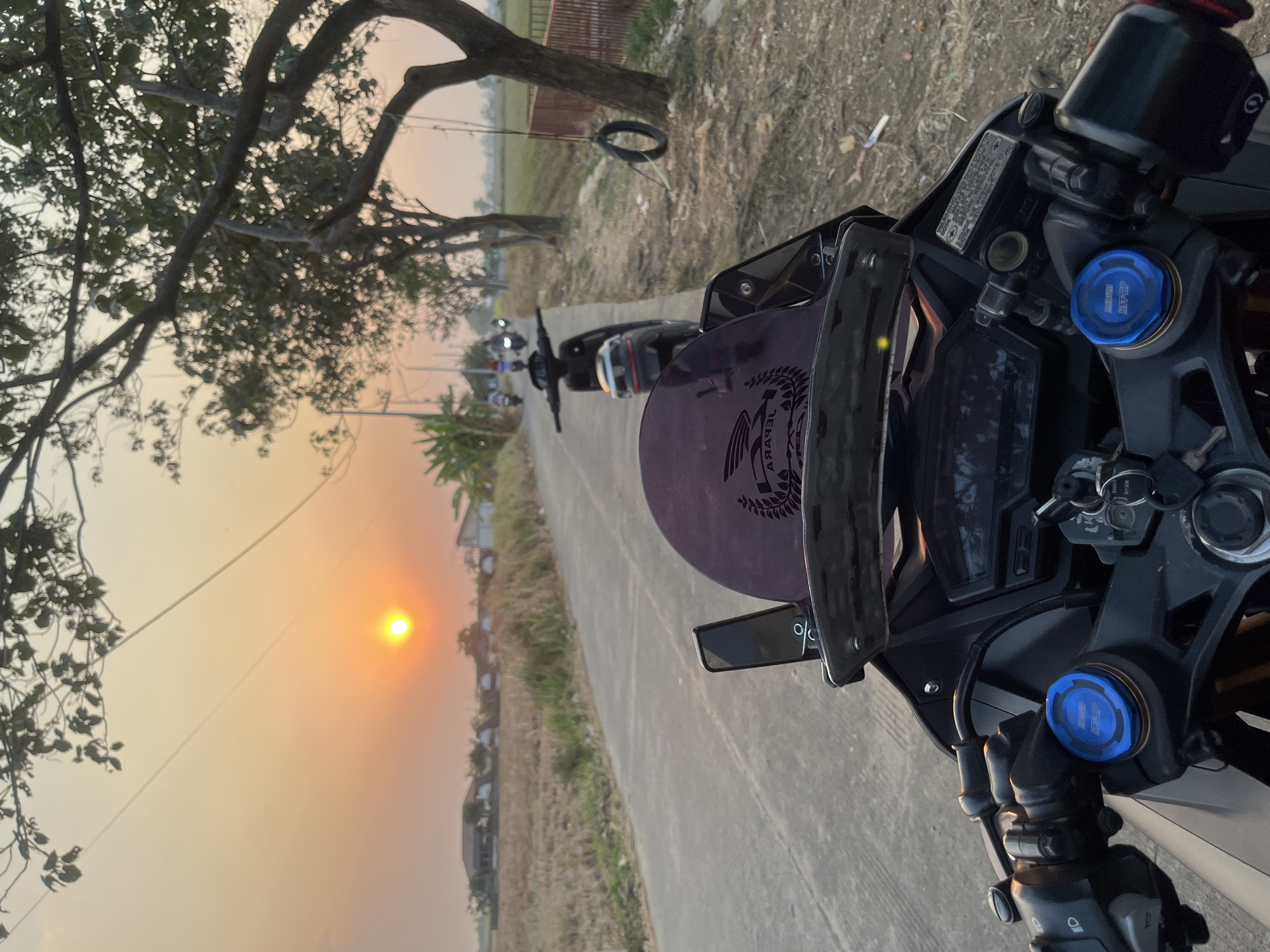 The image captures a sunset scene with the sun low on the horizon, casting a warm orange glow over a rural landscape. The perspective is from the rider of a motorcycle, with the handlebars and part of the dashboard visible in the foreground. The motorcycle appears to be black with blue accents on the front suspension. Behind the motorcycle, a paved road stretches into the distance, lined with trees and dry vegetation. Several other motorcycles are seen further down the road, suggesting a group ride. In the background, there are houses and fields, indicative of a village or rural area. A tire swing hangs from a tree on the right side of the road. The overall atmosphere is serene and peaceful, with the natural beauty of the sunset dominating the scene. The location is identified as Troso, Indonesia.