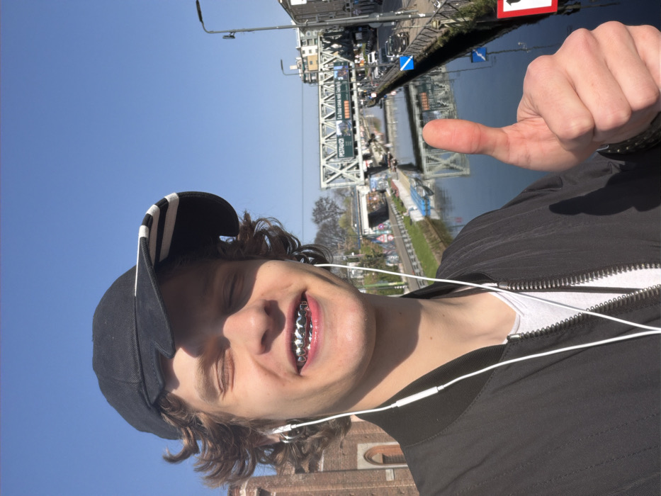 A young individual is captured in a sunlit outdoor selfie, positioned against a clear blue sky. The person, with curly brown hair partially covered by a black baseball cap featuring white stripes on the brim, has their eyes closed and a wide smile, displaying silver grillz on their upper teeth. White wired earbuds are visible, connected to their ears and draped over a black bomber jacket worn over a white t-shirt. Their left hand, prominent in the lower right foreground, displays a thumbs-up gesture.
The background features a canal with calm, reflective water. A substantial metal truss bridge, likely a railway overpass, spans the canal in the midground, carrying an advertising banner that includes the word "PESTAVECCHIA." Roads flank both sides of the canal, with multiple parked cars visible along the right bank. On the far left, a brick building with arched windows is partially visible. Streetlights, trees, and additional urban structures are discernible in the distance. This scene is located in the Navigli district of Milan, Italy.