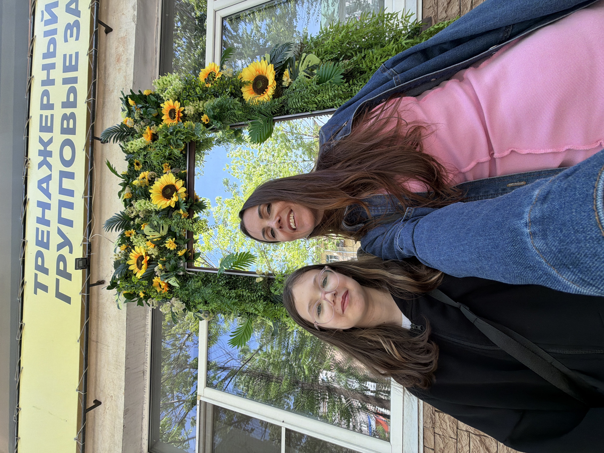 Two women pose for a selfie in front of a building facade. The woman at the top, closer to the camera, wears a pink shirt and denim jacket. She has long, brown hair and is smiling. The woman at the bottom wears glasses and a black jacket. Her long, brown hair is parted on the side.
Behind the women is a window framed with artificial sunflowers and greenery. The window reflects trees and the sky, suggesting it is daytime. To the left of the window is a yellow sign with text in Russian: "ТРЕНАЖЕРНЫЙ ГРУППОВЫЕ ЗА".
The exterior of the building is visible, with brickwork and painted surfaces. Based on the Russian text, the photo was taken in a Russian-speaking area. Birobidzhan, Russia is the only location provided.