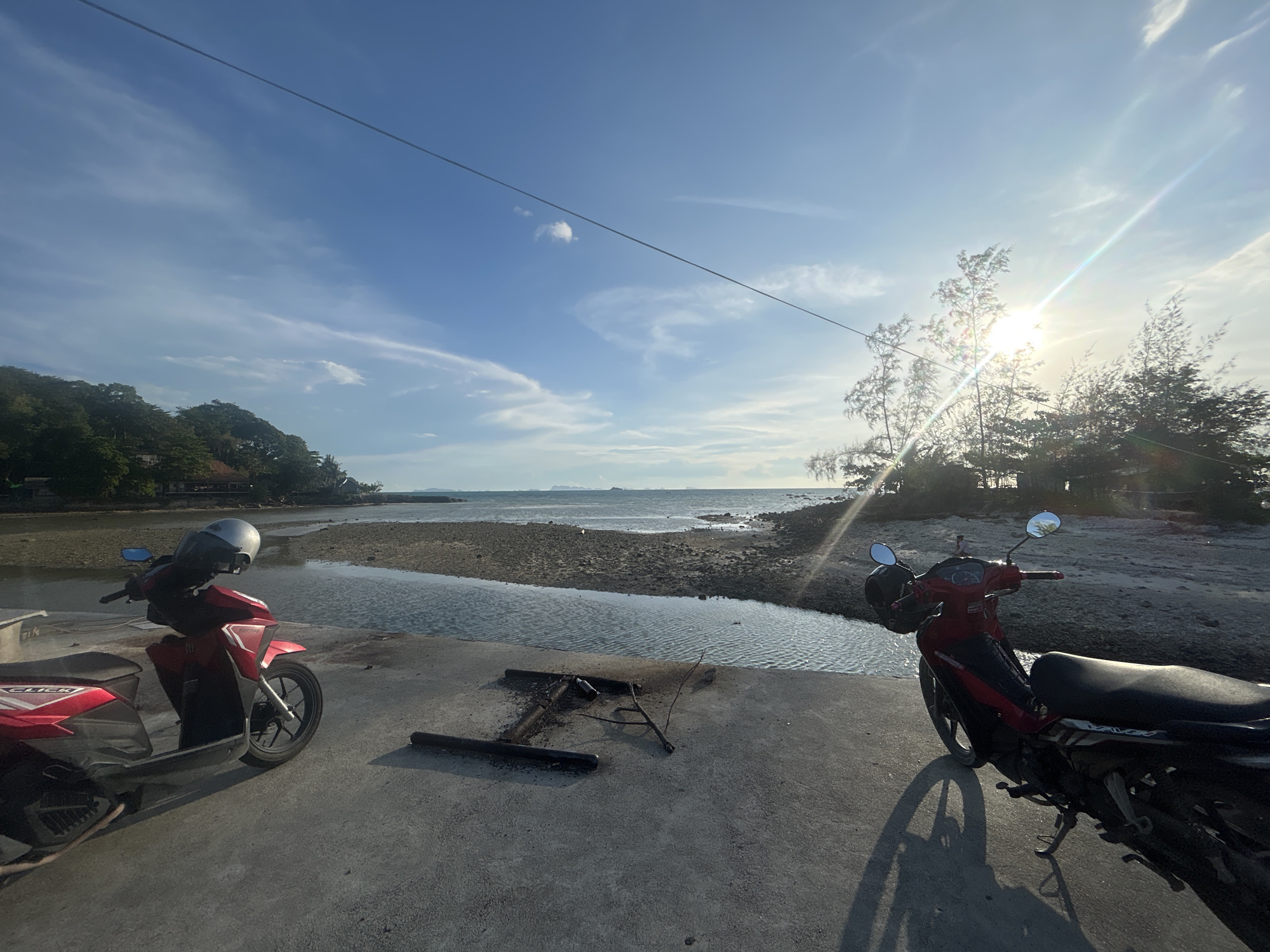 The image depicts a waterfront scene in ตำบลเกาะพะงัน, Thailand. Two red motorbikes are parked on a concrete surface in the foreground. Between them, metal debris and a small branch rest on the concrete. A body of water, possibly a tidal creek or shallow inlet, stretches towards the ocean in the middle ground. The water level is low, exposing a rocky shoreline. In the background, a small island or peninsula covered in trees is visible on the right, and the mainland coastline is on the left. The sky is blue with scattered clouds, and the sun is shining brightly behind the trees on the right, creating a lens flare effect. A power line crosses the upper part of the frame.