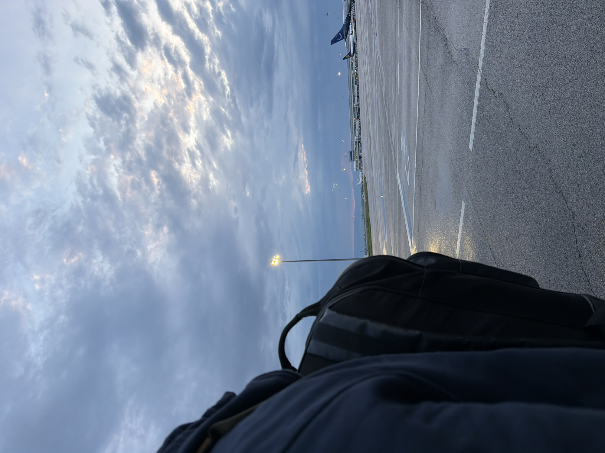 This image captures an airport tarmac in Sofia, Bulgaria, during either dawn or dusk. The sky is largely overcast with dramatic, textured clouds, some of which are subtly illuminated with soft, warm golden and pink light from the sun below the horizon.
In the immediate foreground, a dark-colored bag, resembling a backpack or duffel bag, is prominently positioned, alongside a portion of dark clothing, indicating the perspective of a traveler. A tall light pole with a glowing yellow lamp provides artificial light against the darkening or brightening sky.
In the mid-ground, a commercial aircraft is parked on the asphalt tarmac. Its tail displays a blue and white livery with the airline's name, "RYANAIR," clearly visible. White painted lines on the tarmac mark out designated areas, and some minor cracks are visible on the asphalt surface. The overall scene conveys a quiet, atmospheric moment at an airport, likely before a departure or after an arrival, with the low light and artificial illumination creating a sense of transition.