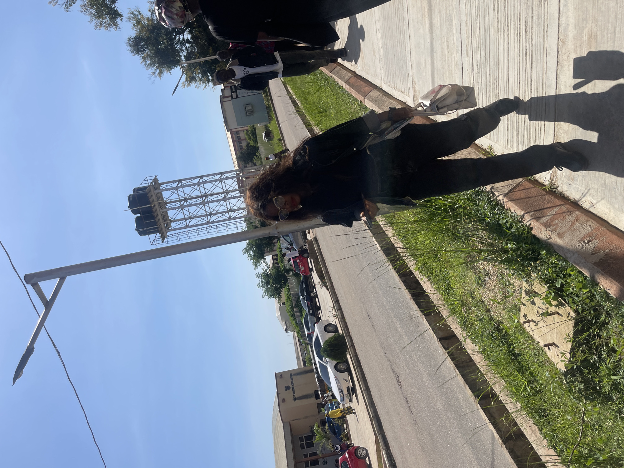 The image depicts an outdoor scene in a city, likely Rafin Par, Nigeria, under a clear blue sky. In the foreground, a woman with long, dark, wavy hair and glasses is walking on a concrete sidewalk. She is wearing a dark jacket and dark pants, and carrying a light-colored handbag. Her right leg is bent, and she appears to be in motion. Behind her, a shadow of her hand suggests she might be making a peace sign or waving.
Several other individuals are walking in the background on the same sidewalk, including a man in a white t-shirt with a logo and dark pants, and another person in dark clothing. Further back, a street runs parallel to the sidewalk, with several cars parked along its edge. A white sedan is prominently visible, along with other vehicles in various colors.
On the left side of the image, a building with "FACULTY OF MANAGEMENT SCIENCES" visible on its facade stands beside the street. Beyond the building, a tall metal tower with water tanks is situated, characteristic of urban infrastructure. The environment includes green grass growing along the edge of the sidewalk and road. The overall impression is of a sunny day on a campus or a public area within a Nigerian city.